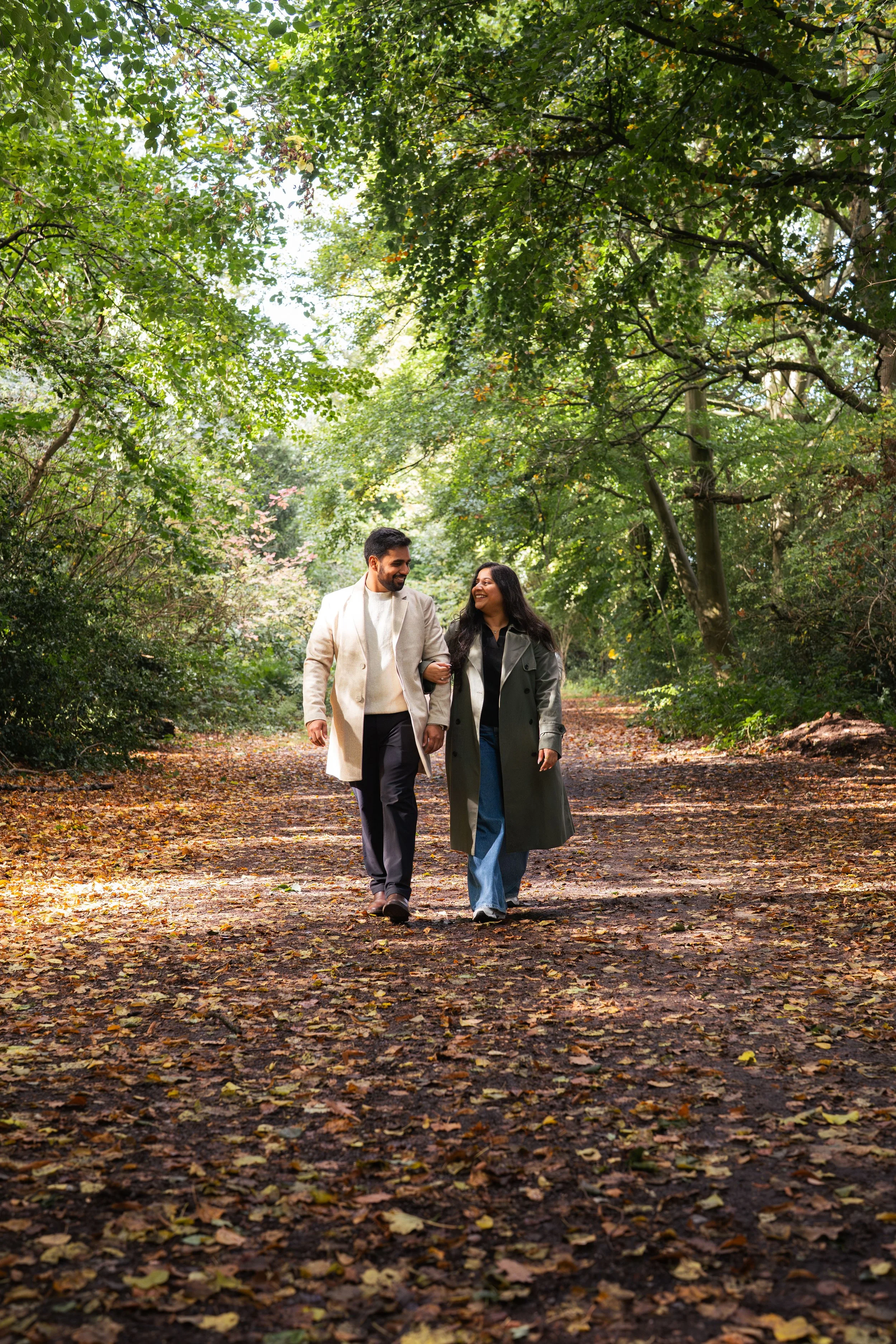 A man and woman walking arm in arm on a fallen leaf-covered path through a lush green wooded area, smiling and looking at each other.