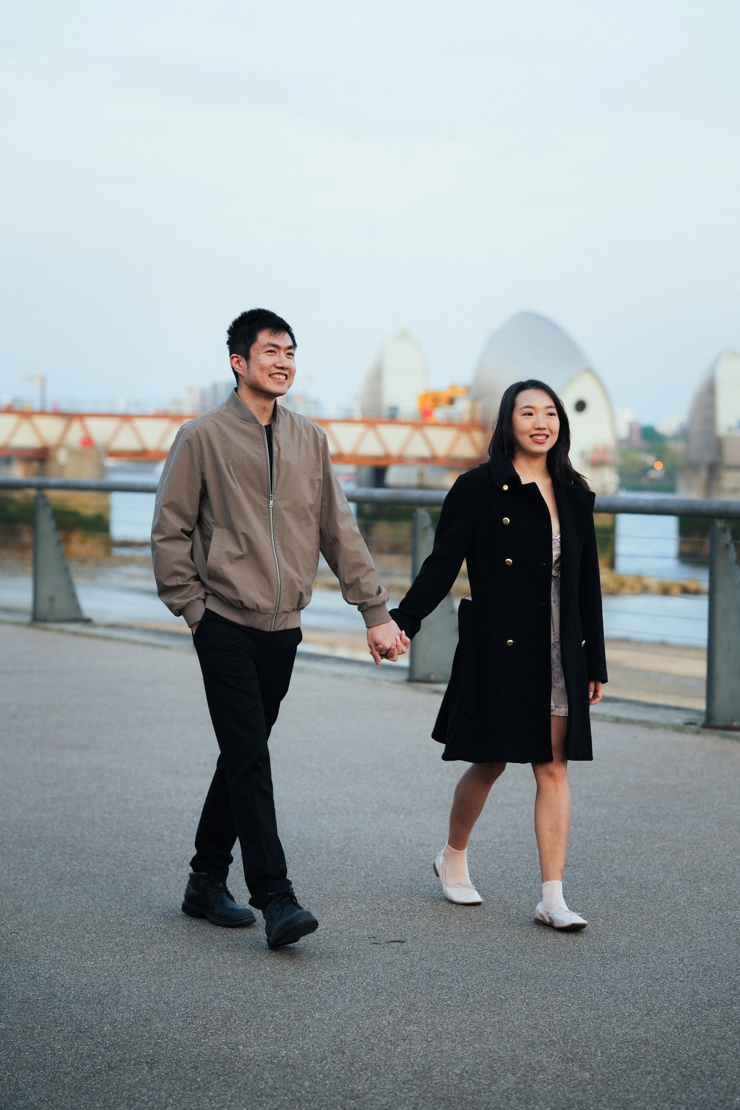 A young couple, holding hands and smiling, walking along a waterfront promenade during the evening. The city skyline and a bridge are visible in the background.