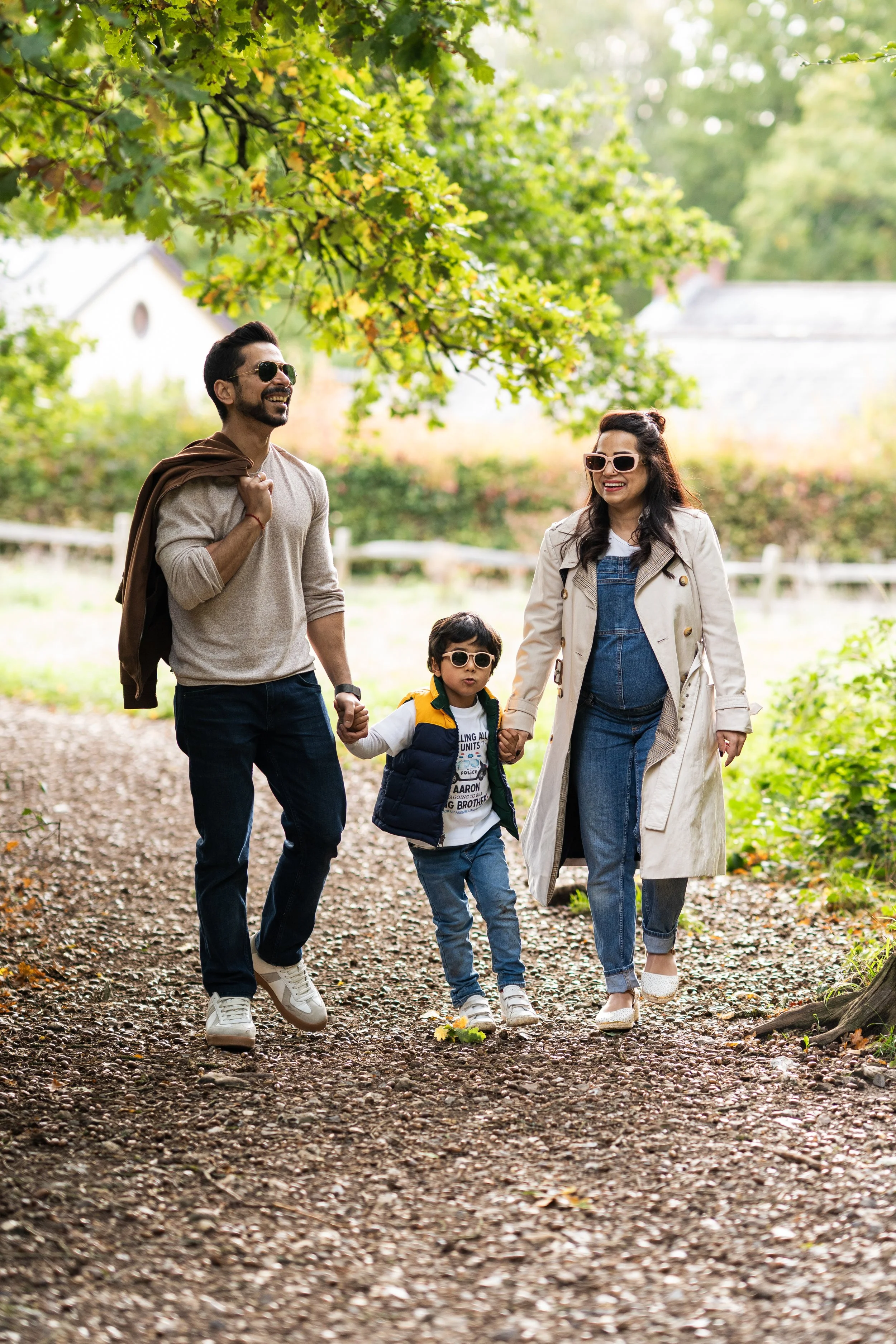 A happy family of three, a man, woman, and young boy, walking hand-in-hand through a park with trees and greenery on a bright day. They are all wearing sunglasses and casual clothing.