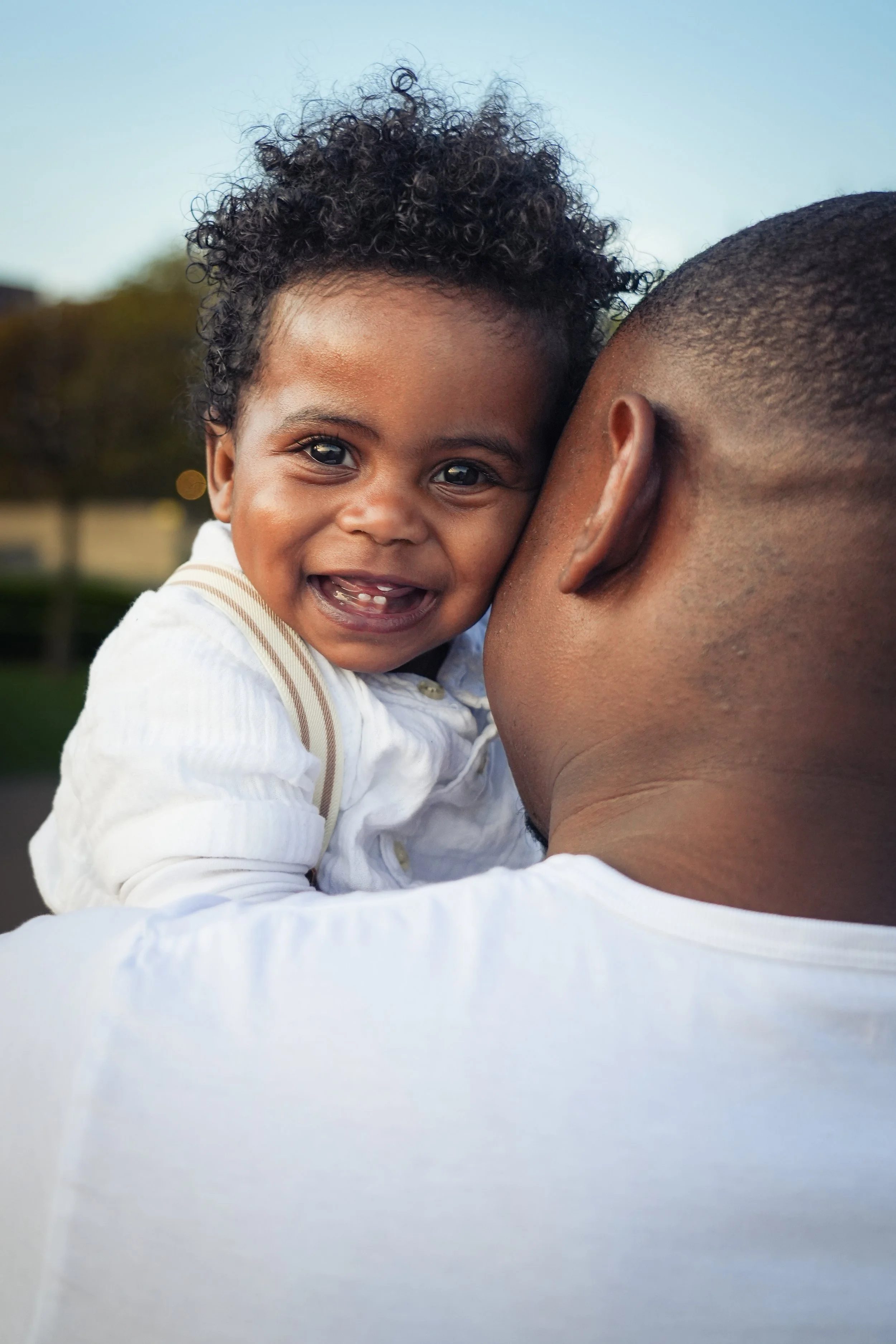 A young child with curly hair smiling, being held by an adult man with close-cropped hair outdoors during daytime.