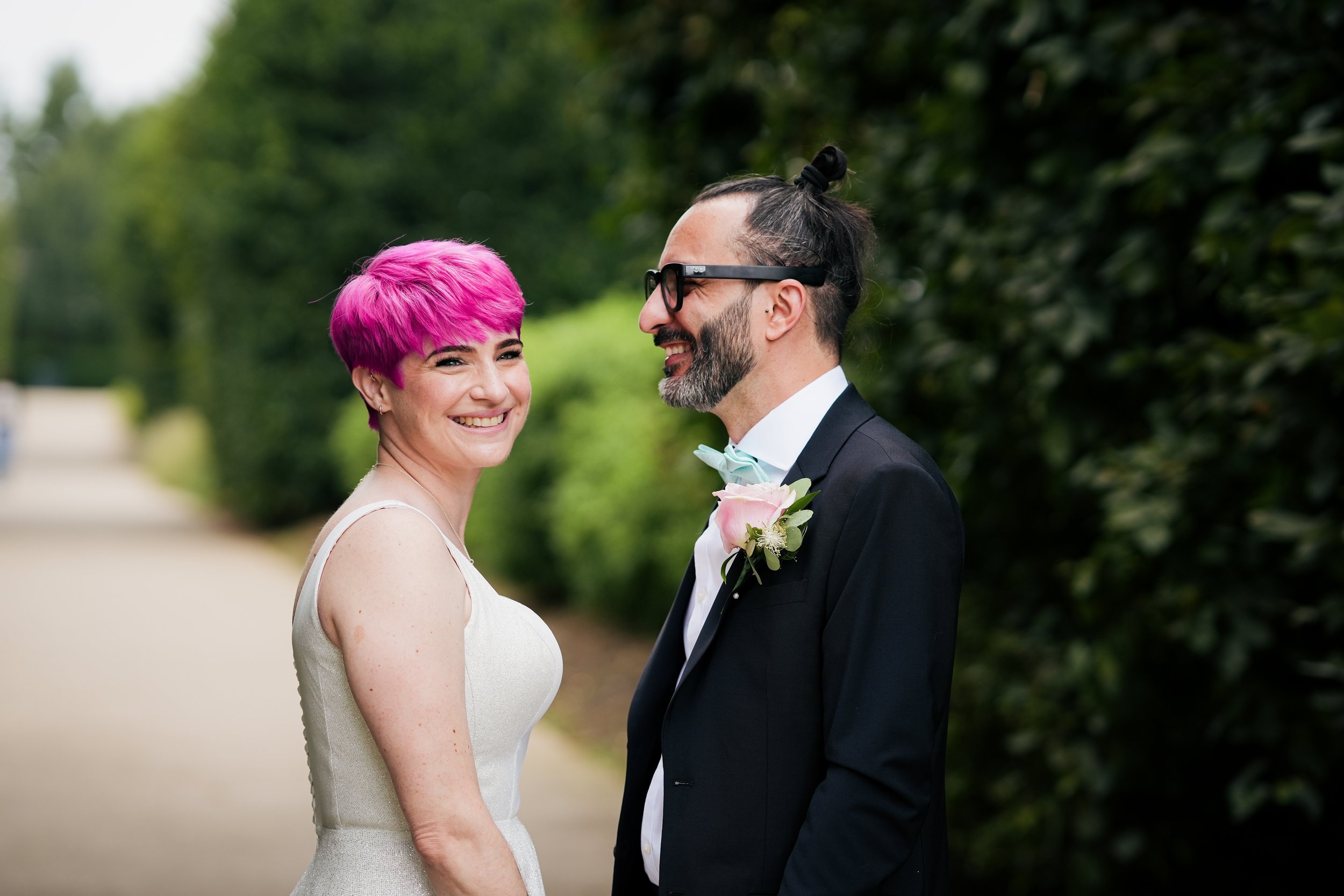 A man and woman smiling at each other outdoors, dressed in wedding attire, with the woman having short bright pink hair and the man wearing glasses, a black suit, and a pink boutonniere.