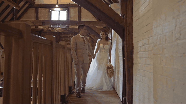 A bride and groom walk hand in hand down a rustic indoor hallway with wooden beams and brick walls, dressed in wedding attire.