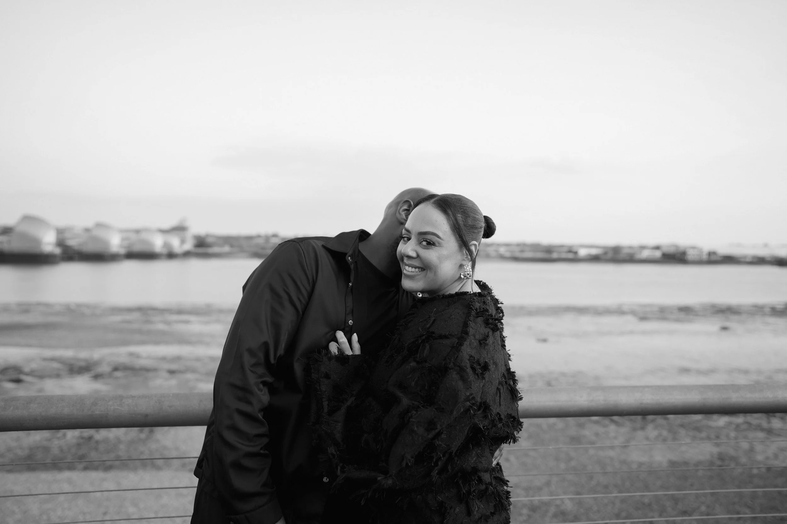 A smiling woman with earrings stands in front of a man who is whispering in her ear near a body of water, with buildings visible in the background. The photo is in black and white.