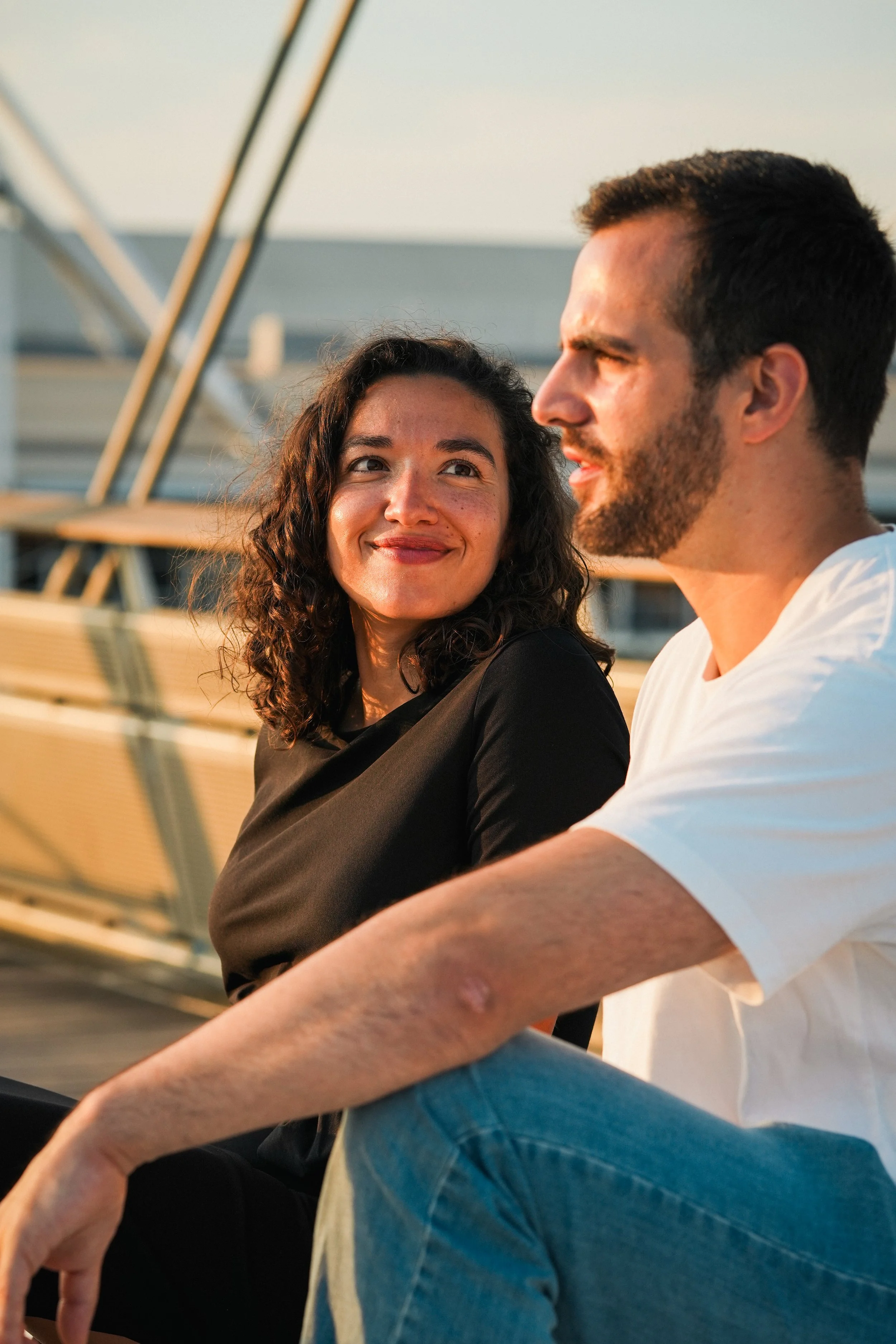 A woman and a man sit on a dock at sunset, looking at each other with smiles.