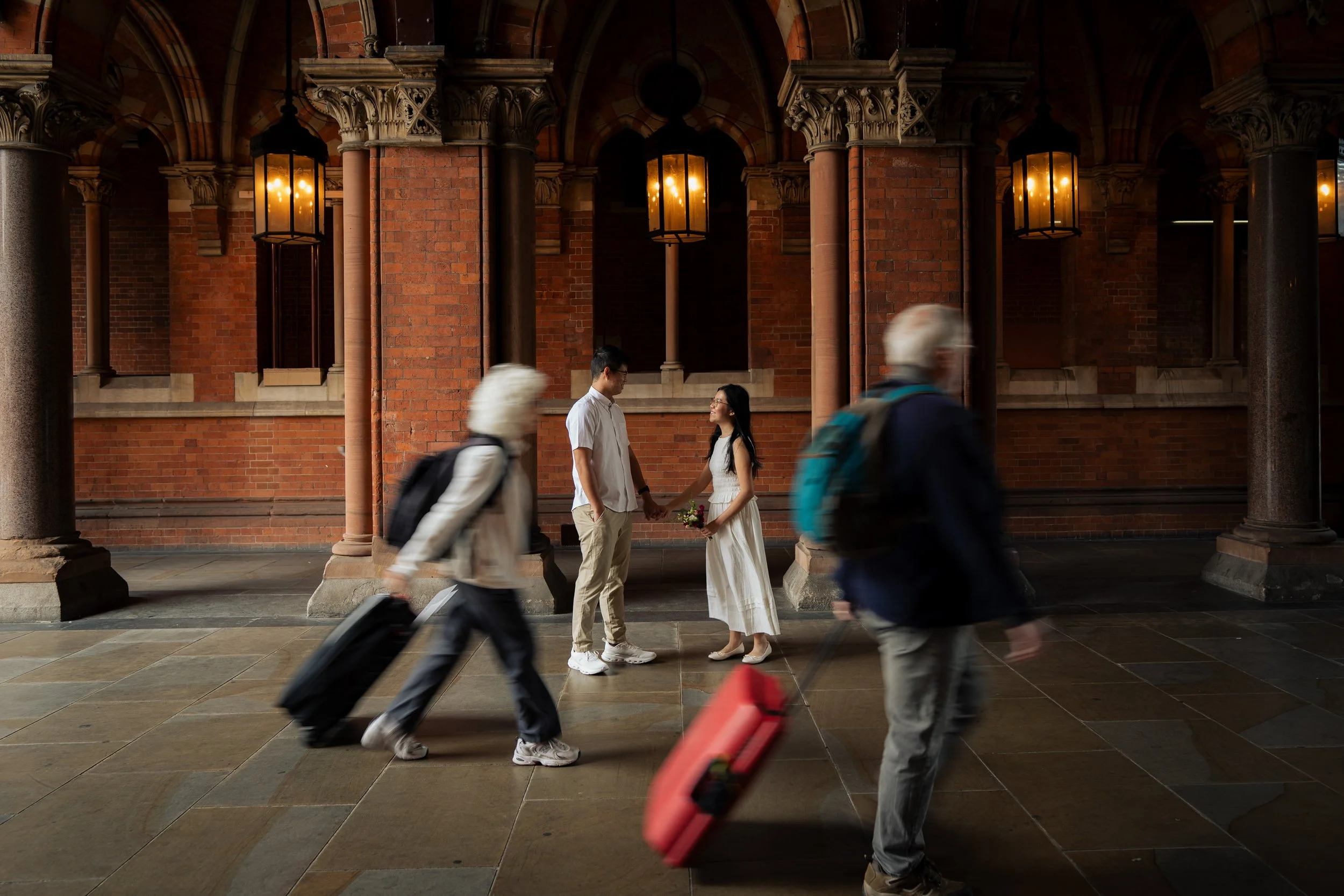 A bride and groom holding hands and facing each other inside a brick building with columns and hanging lanterns, surrounded by blurred travelers with suitcases.