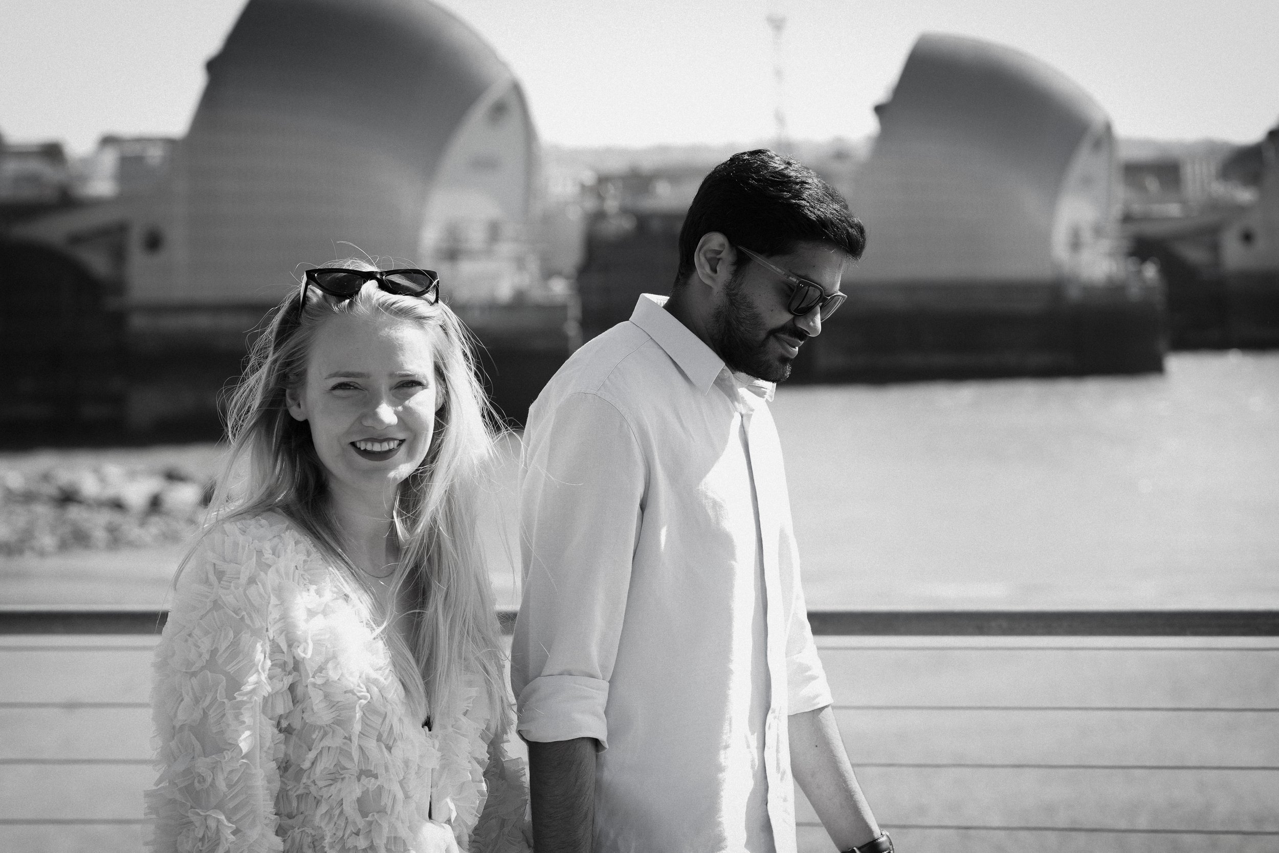 A black and white photo of a smiling woman with long blonde hair and sunglasses on her head, paired with a man with dark hair and beard, wearing sunglasses and a light-colored shirt, walking along a waterfront with large buildings or structures in th