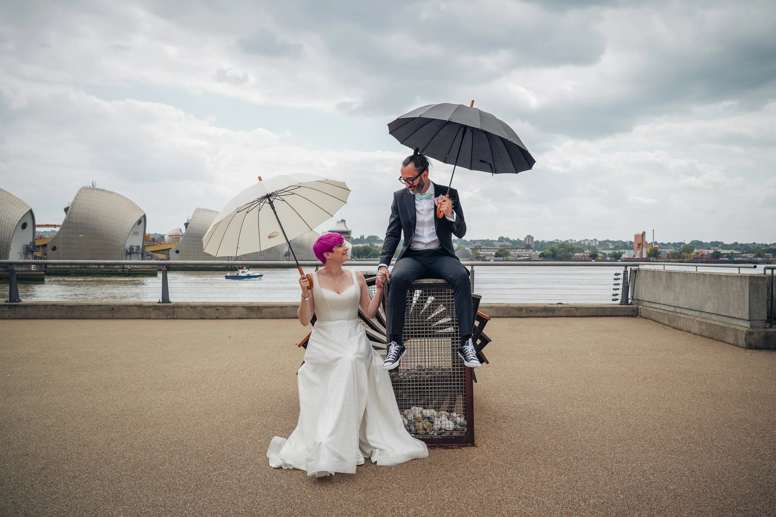 A woman in a wedding dress with bright pink hair holding a white umbrella, sitting on the ground. A man in a tuxedo with glasses and a man bun sitting on a small structure with an umbrella, holding hands and looking at each other. They are outdoors b