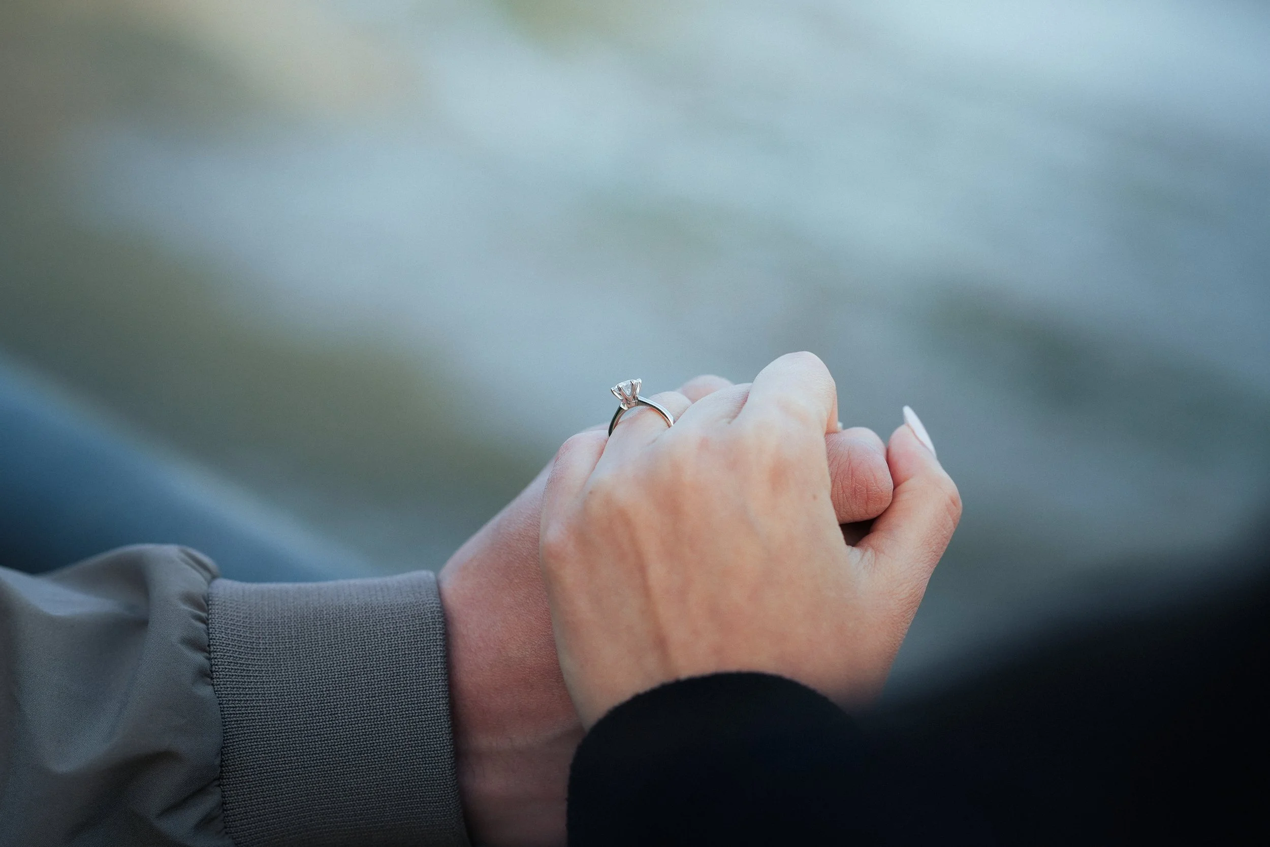Close-up of two people holding hands, one wearing a ring with a diamond, possibly an engagement or wedding ring.