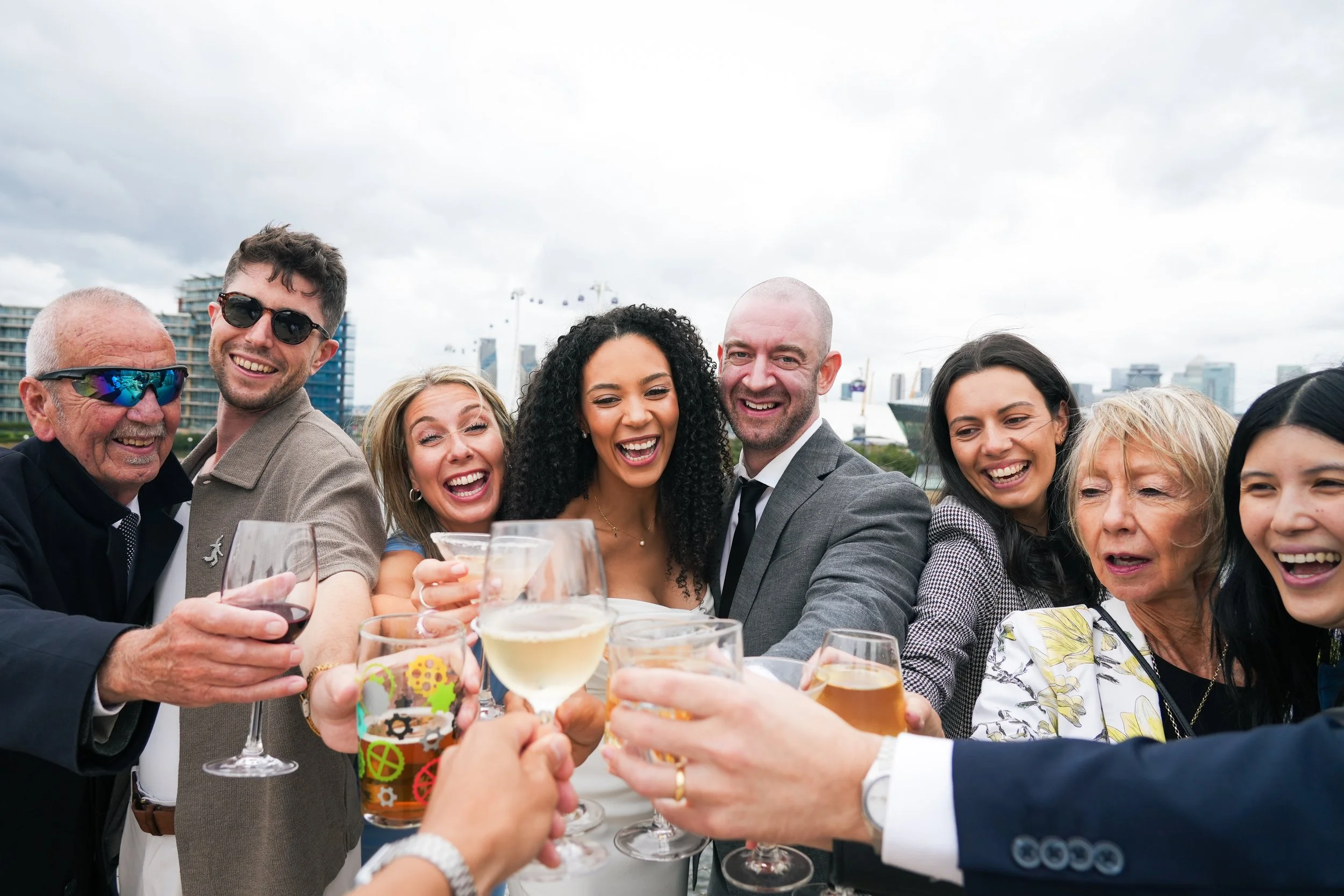 Group of people celebrating outdoors with drinks, smiling and toasting, city skyline in background.