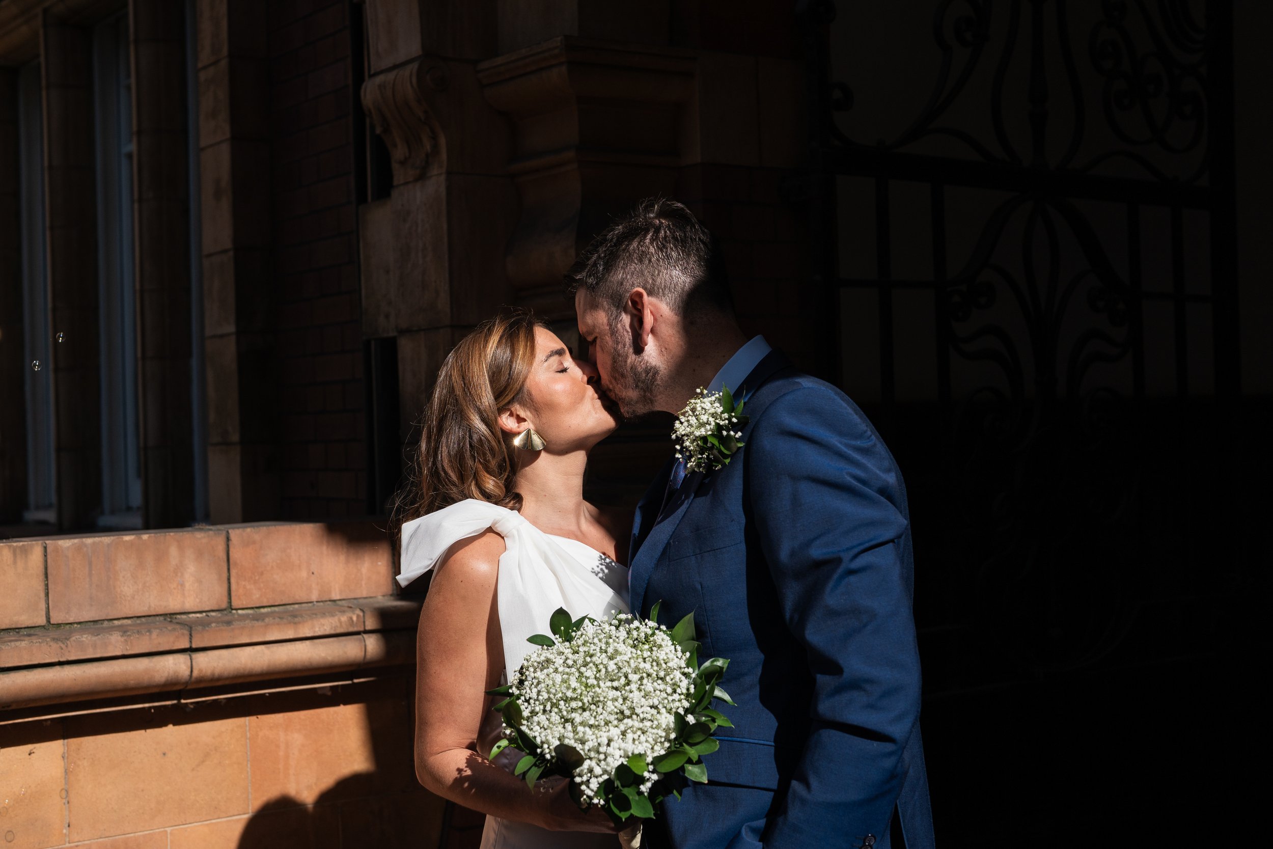 A bride and groom sharing a kiss outdoors during their wedding day, with the bride holding a bouquet of white baby's breath and greenery, and the groom dressed in a blue suit.