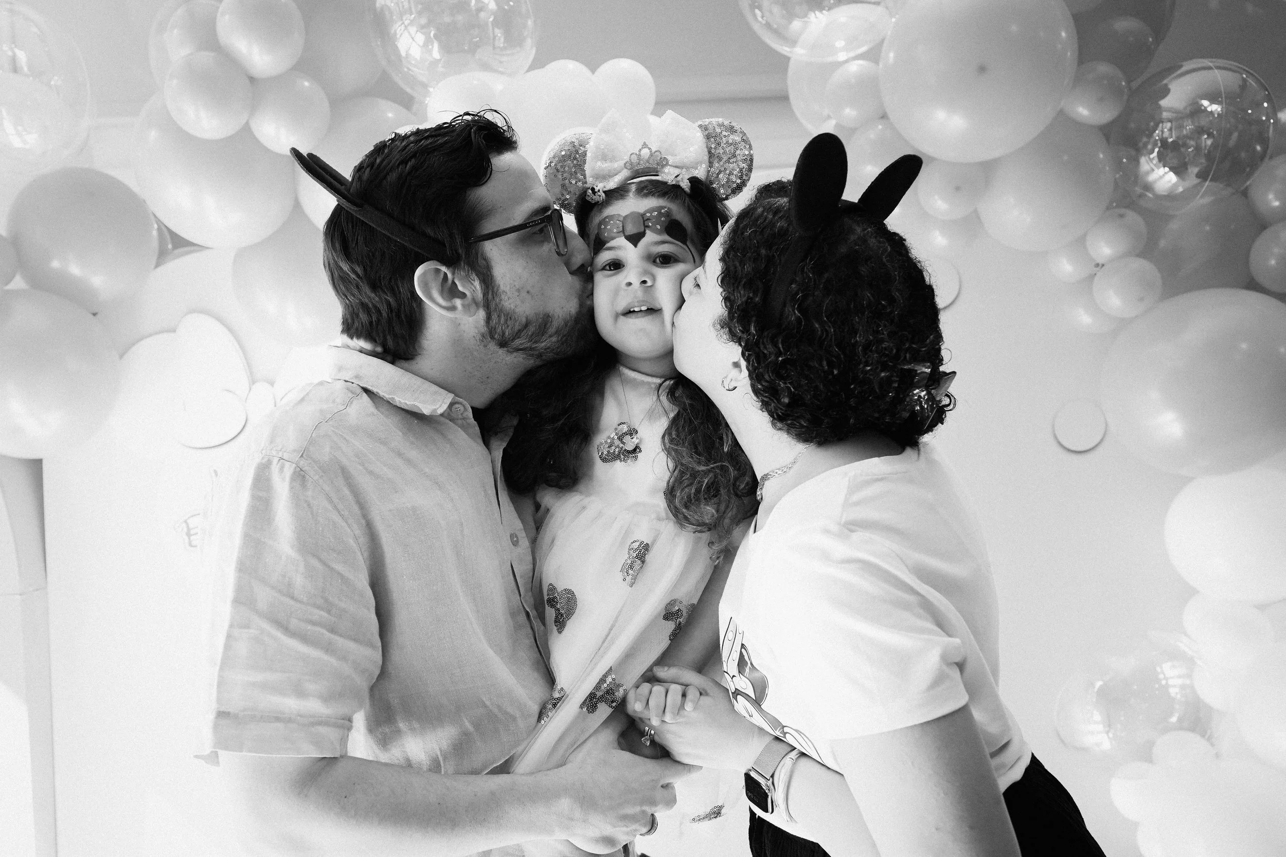 A family of three, with the father and mother giving kisses to their young daughter who is dressed as a princess with a crown and face paint, in a setting decorated with balloons.