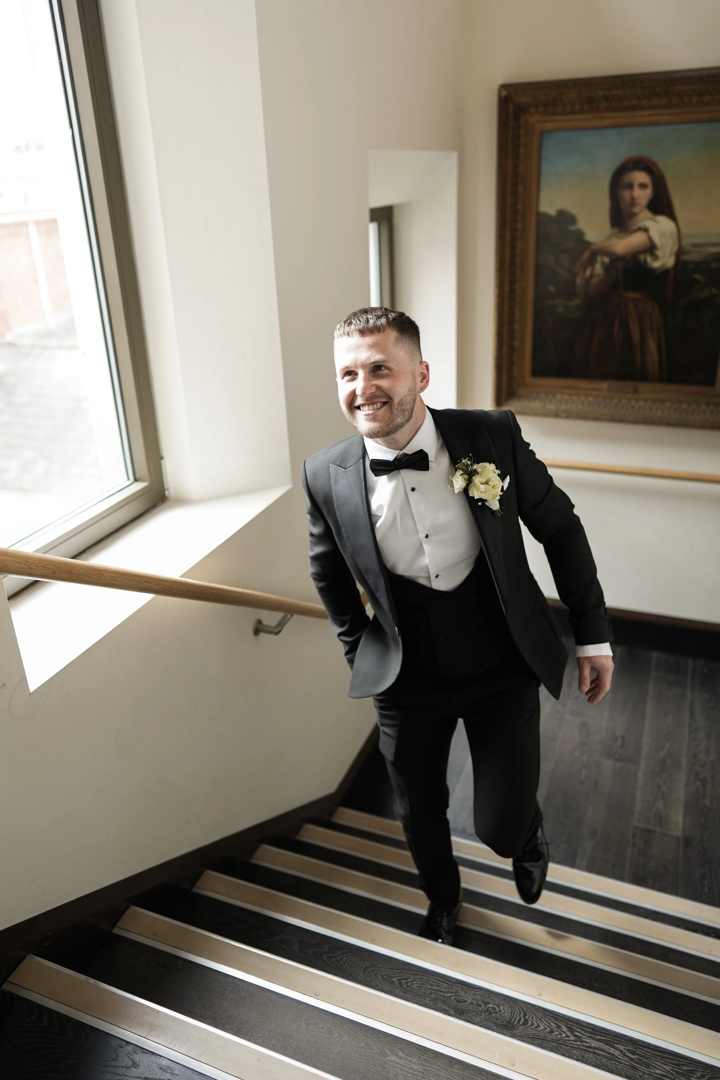 A smiling man in a black tuxedo with a white shirt and black bow tie walking up a staircase, with a flower boutonniere on his lapel, near a window and a framed painting of a woman.
