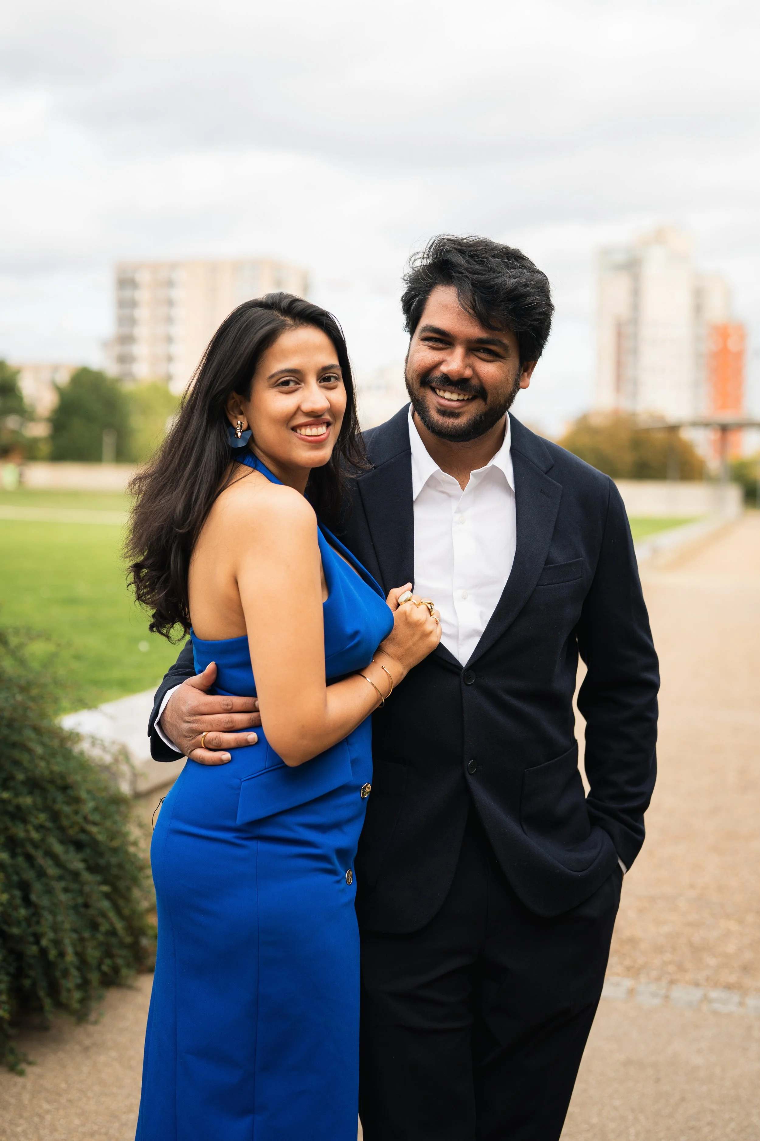 A smiling couple standing outdoors on a cloudy day, with buildings and greenery in the background. The woman is wearing a blue dress and the man is in a dark suit with a white shirt.