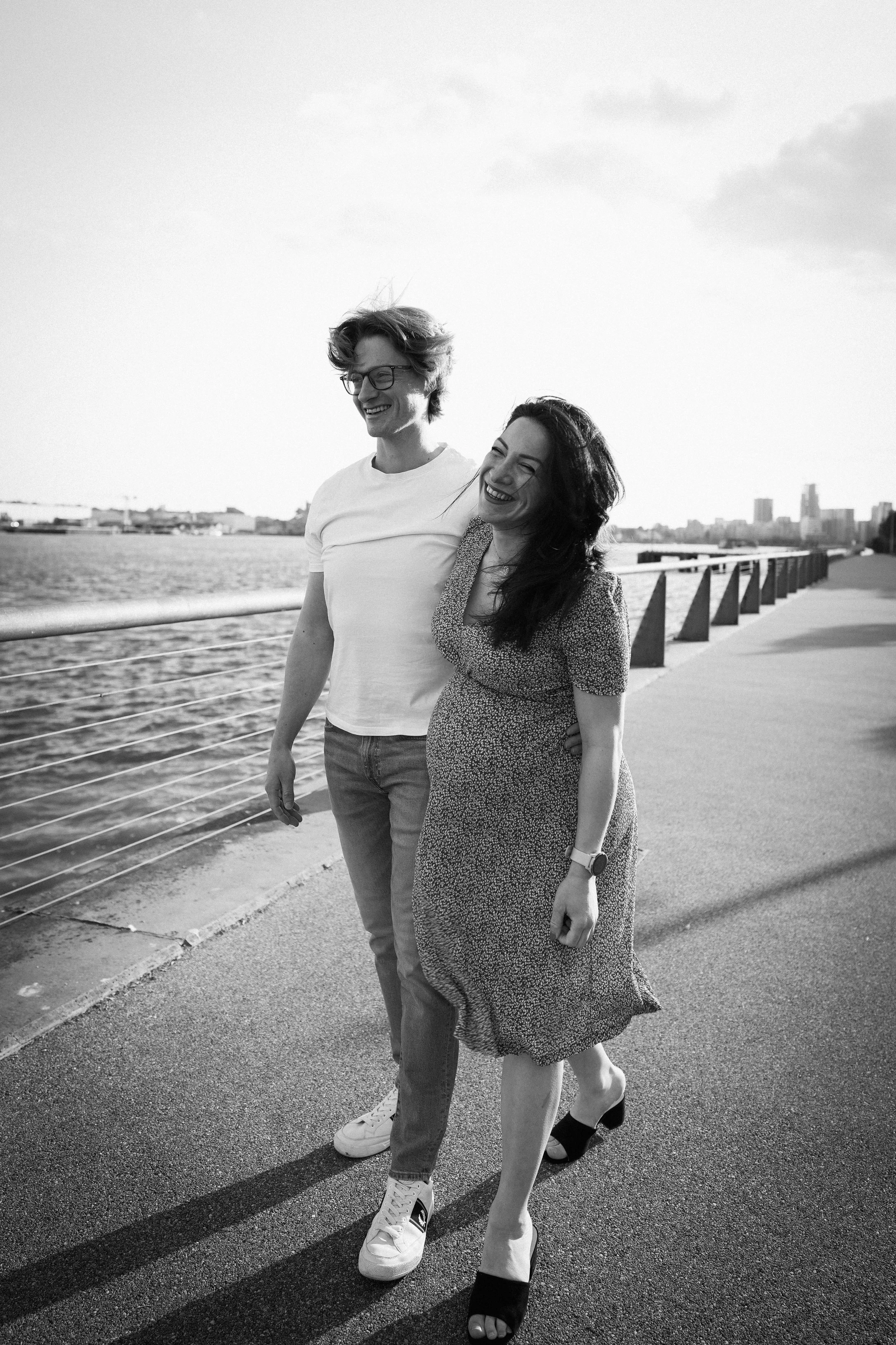 A black and white photo of a smiling couple walking by a river on a sidewalk, with city buildings in the background.