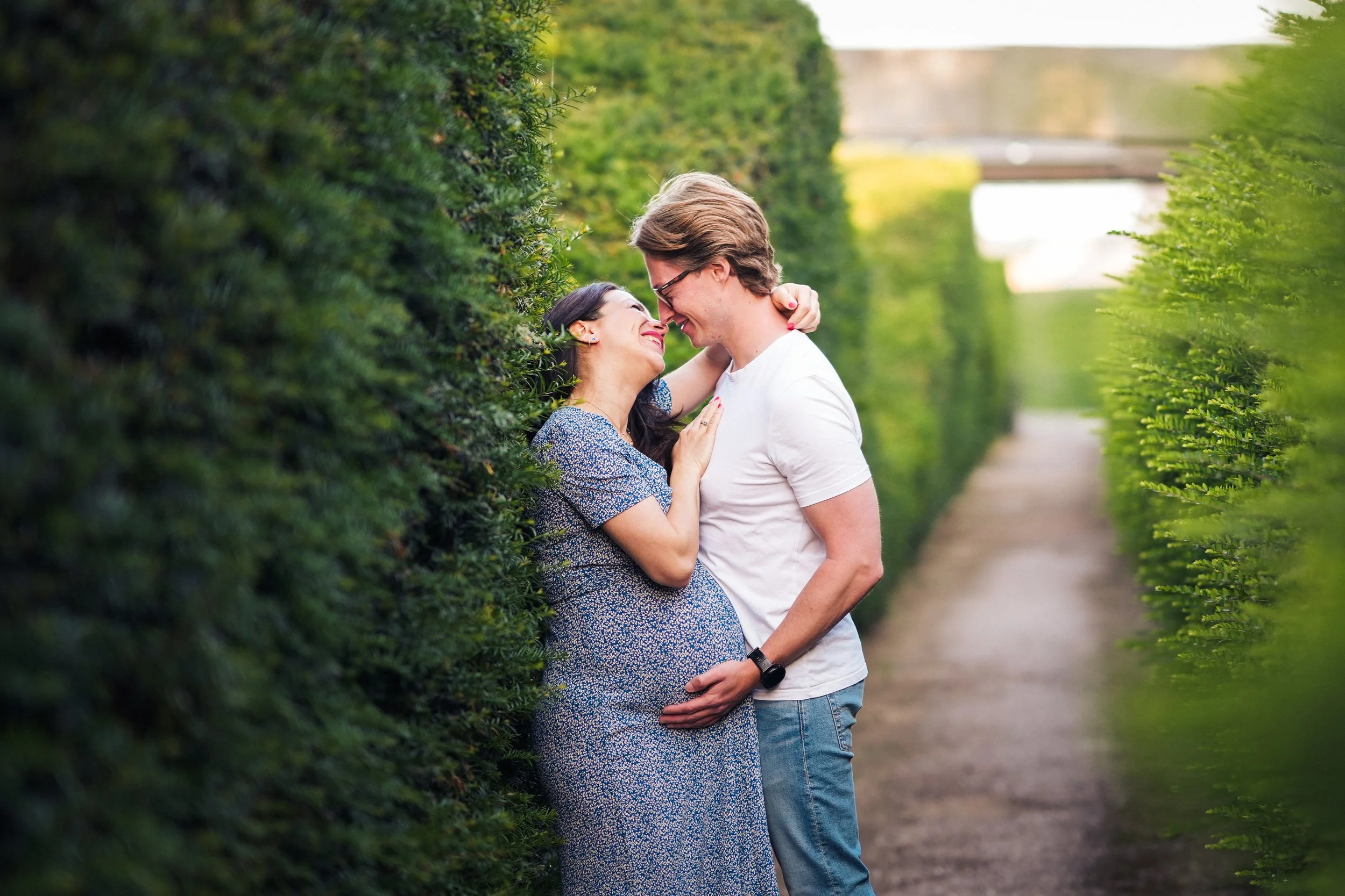 A happy couple is standing in a green hedge maze, embracing and smiling at each other, with the woman touching the man's face and the man touching her pregnant belly, in a lush outdoor setting.