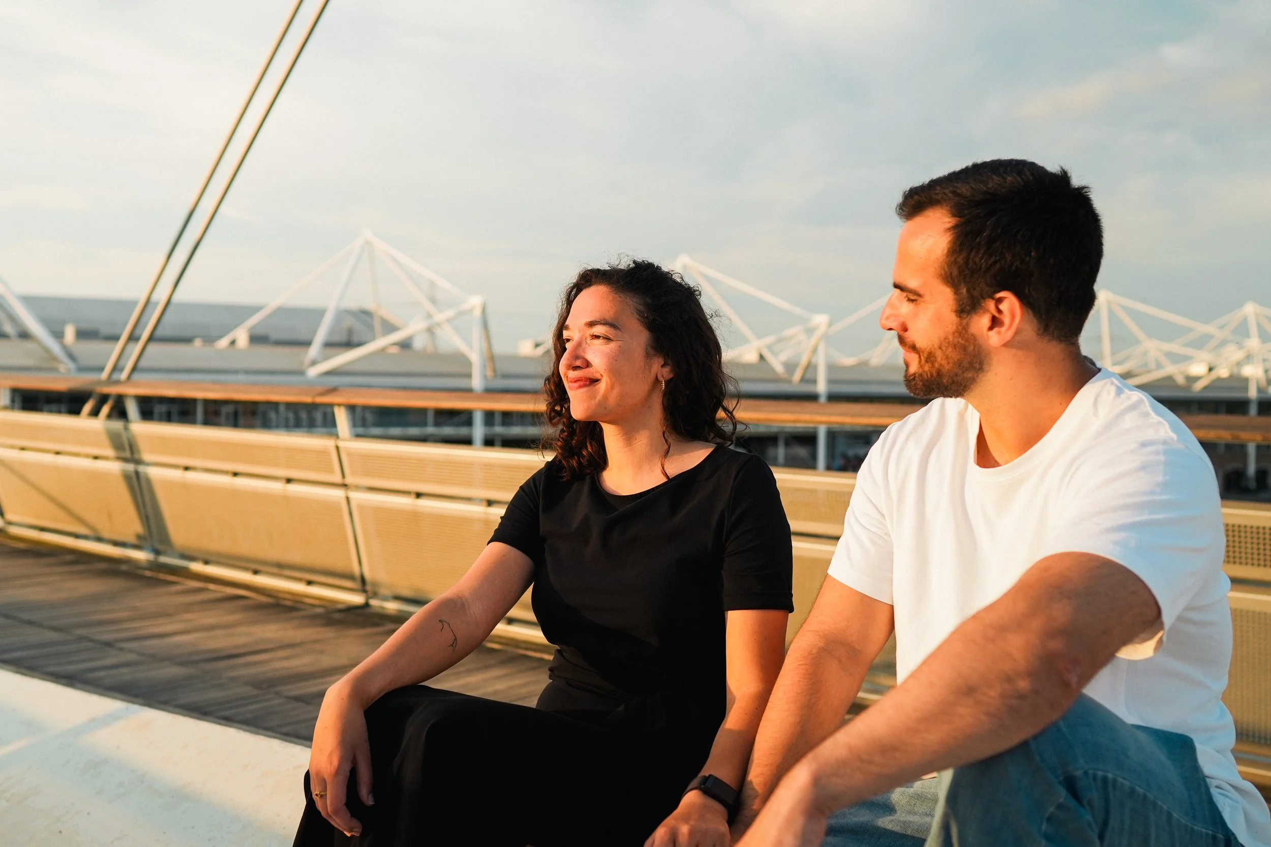 A woman and a man sitting outdoors on a sunny day, looking at each other, with a modern architectural structure in the background.