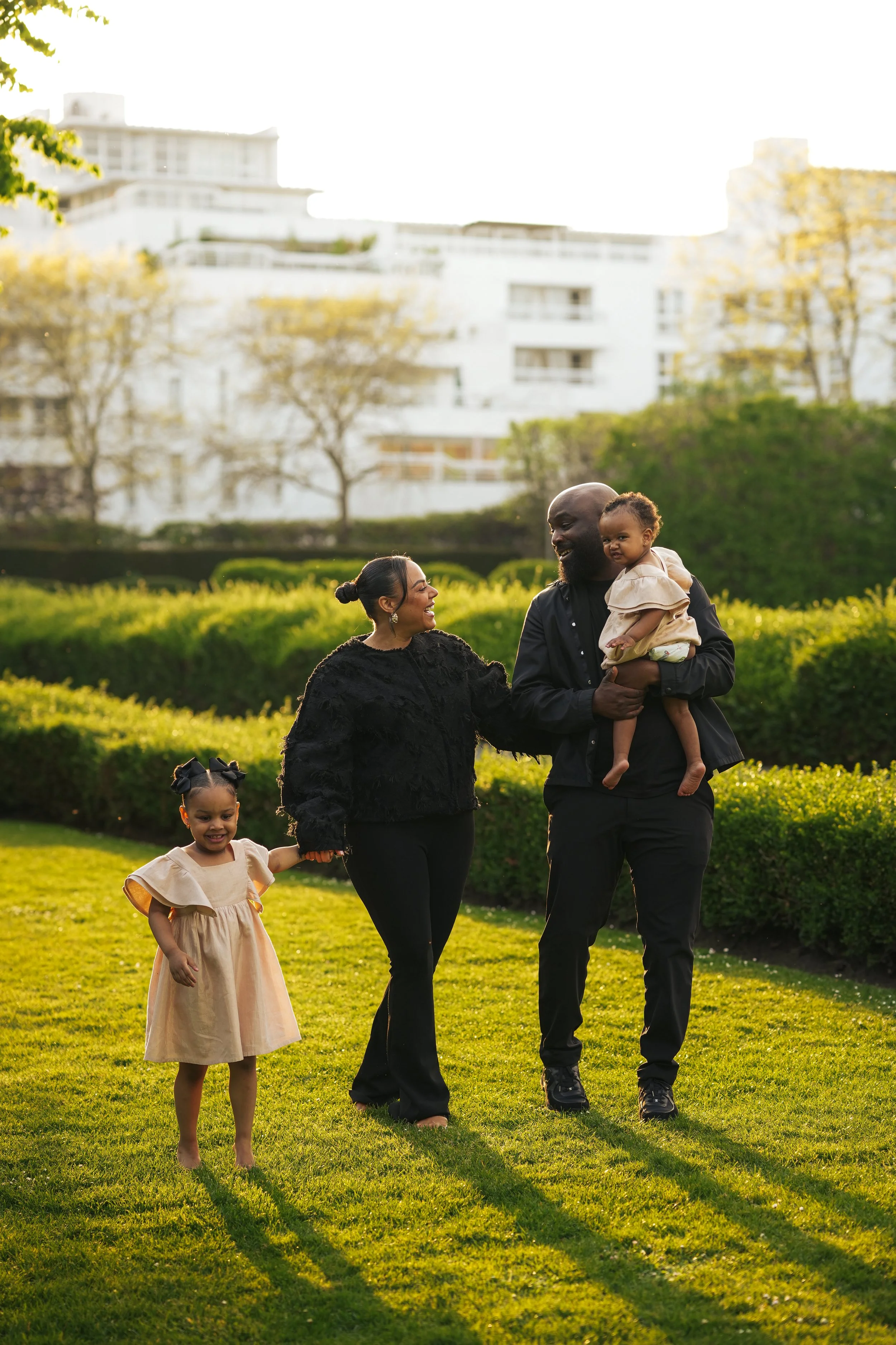 Happy family walking in a park during sunlight, with a woman and a man holding children and a girl walking beside them on a grassy area.