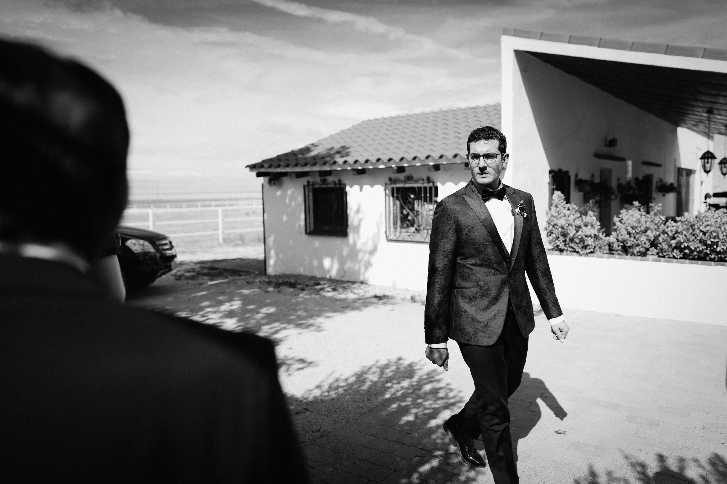 Man in tuxedo walking outdoors near a white house with a tiled roof.