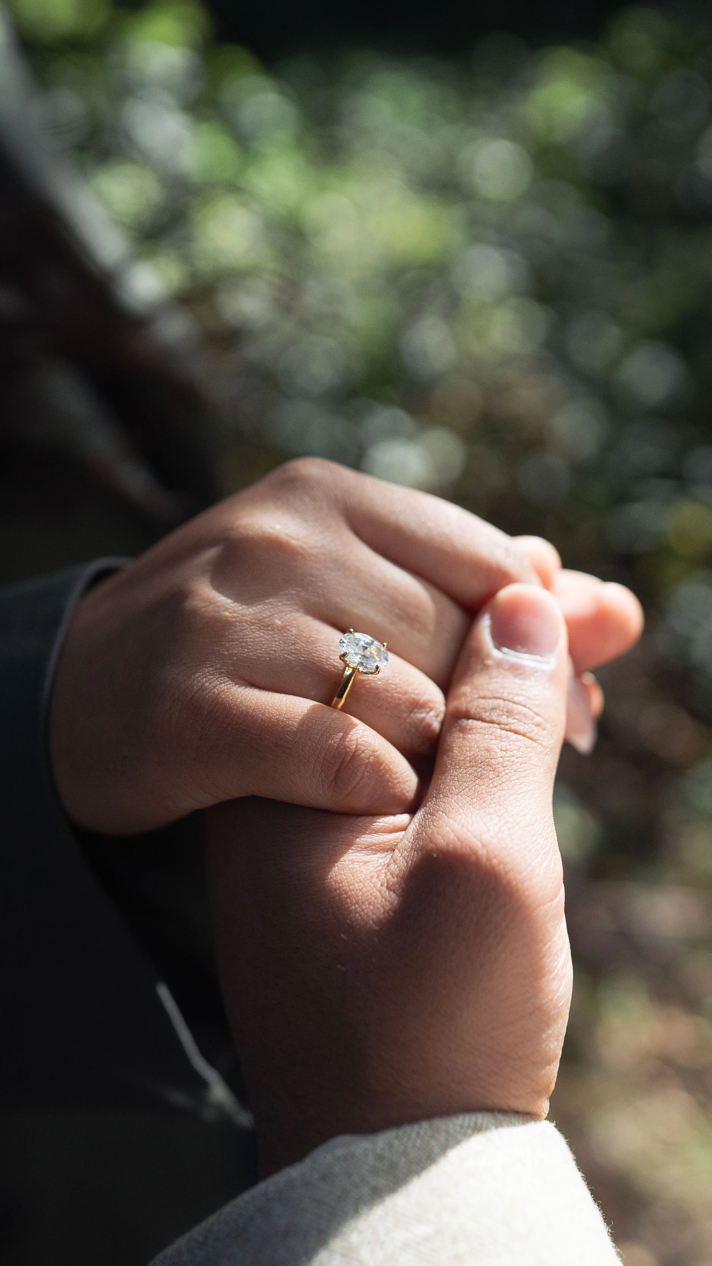 Close-up of a hand with an engagement ring featuring a large round diamond in a prong setting, outdoors with blurred green foliage in the background.