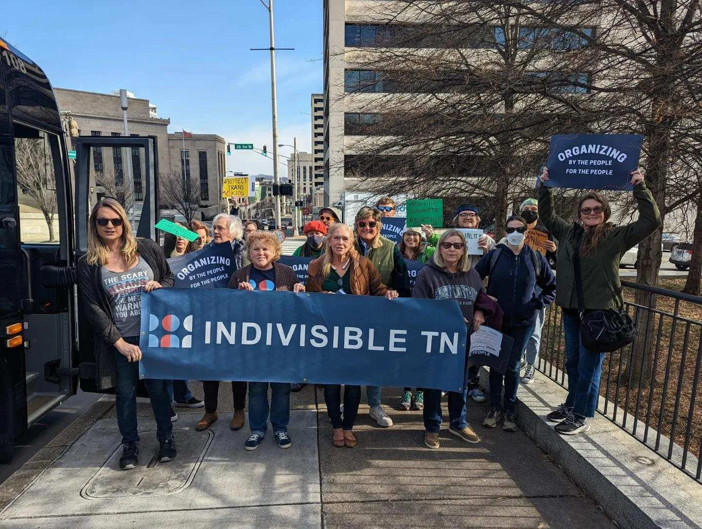 Group of people holding a large blue banner that reads 'INDIVISIBLE TN' at a protest or rally, some holding signs advocating for organizing by the people and protecting trans kids, in an urban area on a sunny day.