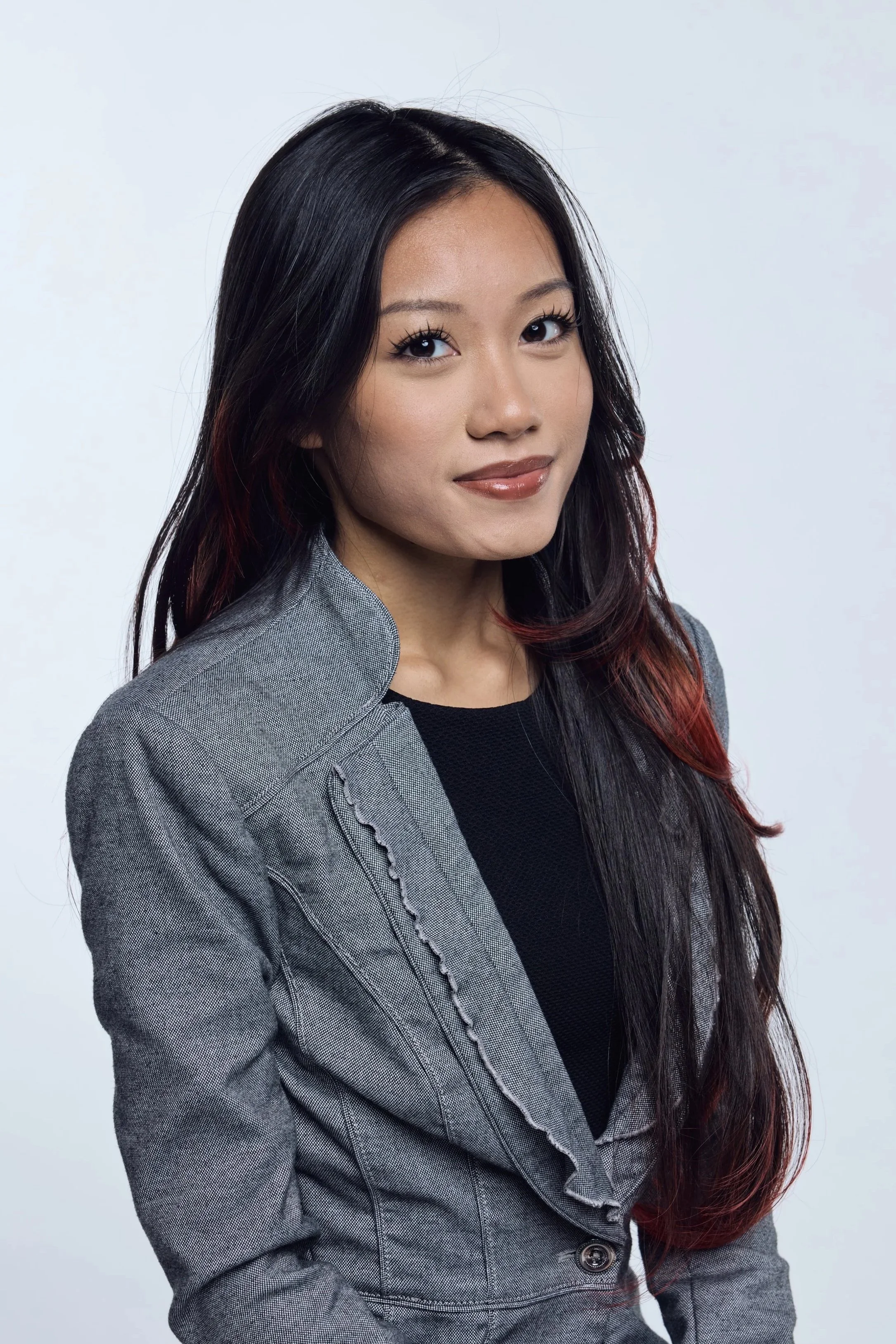 Portrait of a young woman with long black hair and subtle makeup, wearing a gray blazer over a black top, posing against a plain white background.