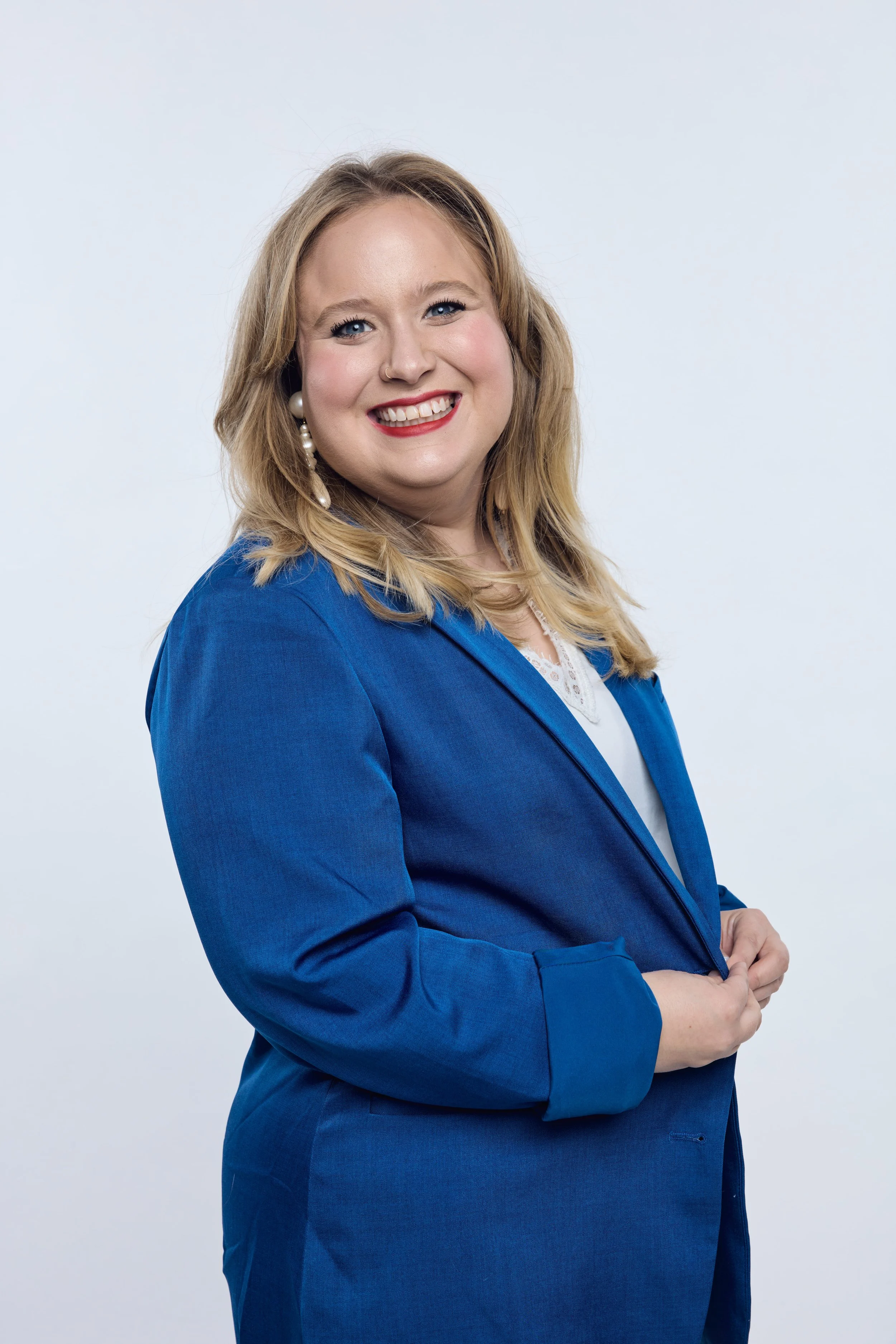 A smiling woman with blonde hair wearing a blue blazer and white top, standing against a plain light gray background.