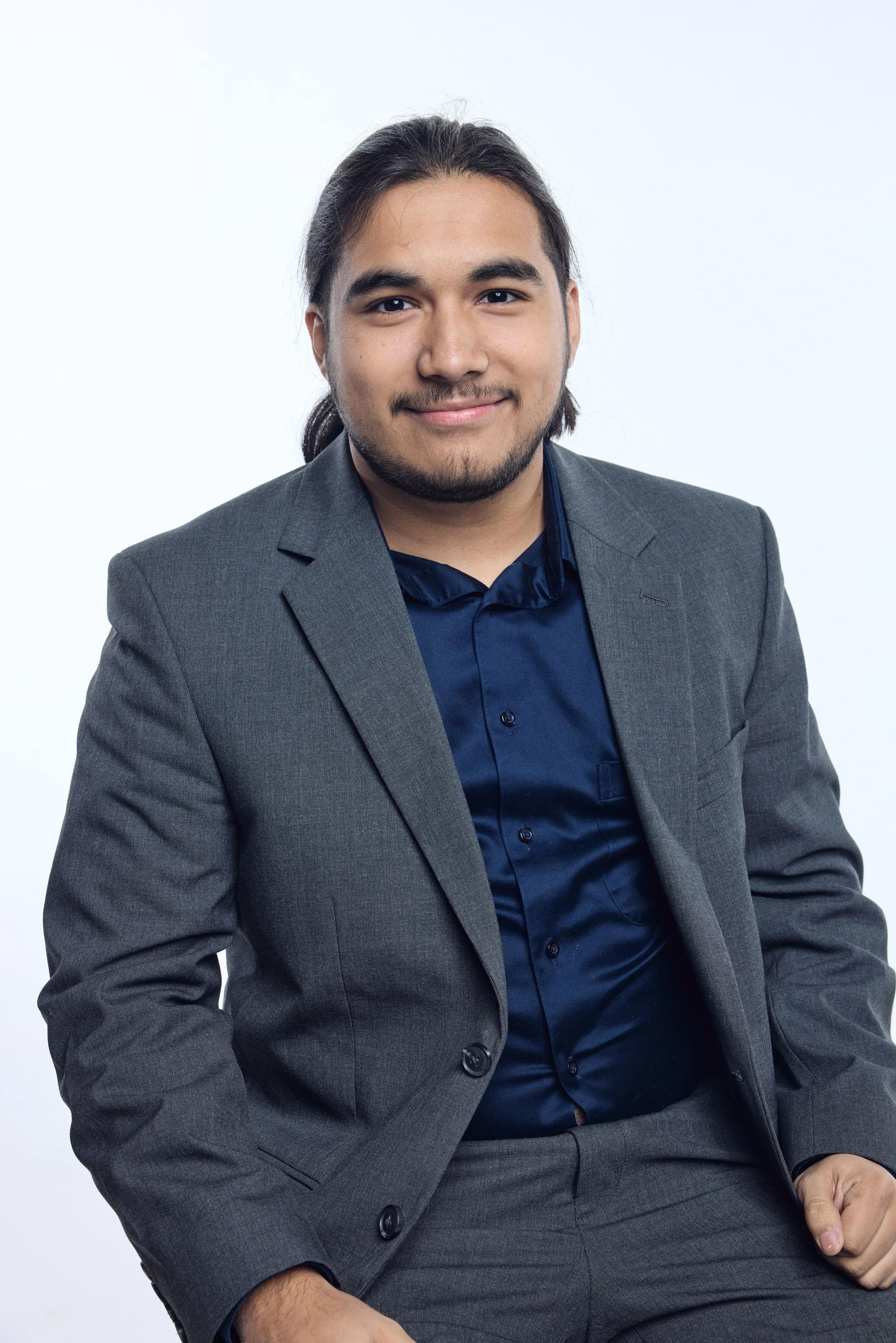 A man with long dark hair, beard, and mustache wearing a gray suit and navy blue shirt, smiling against a plain white background.