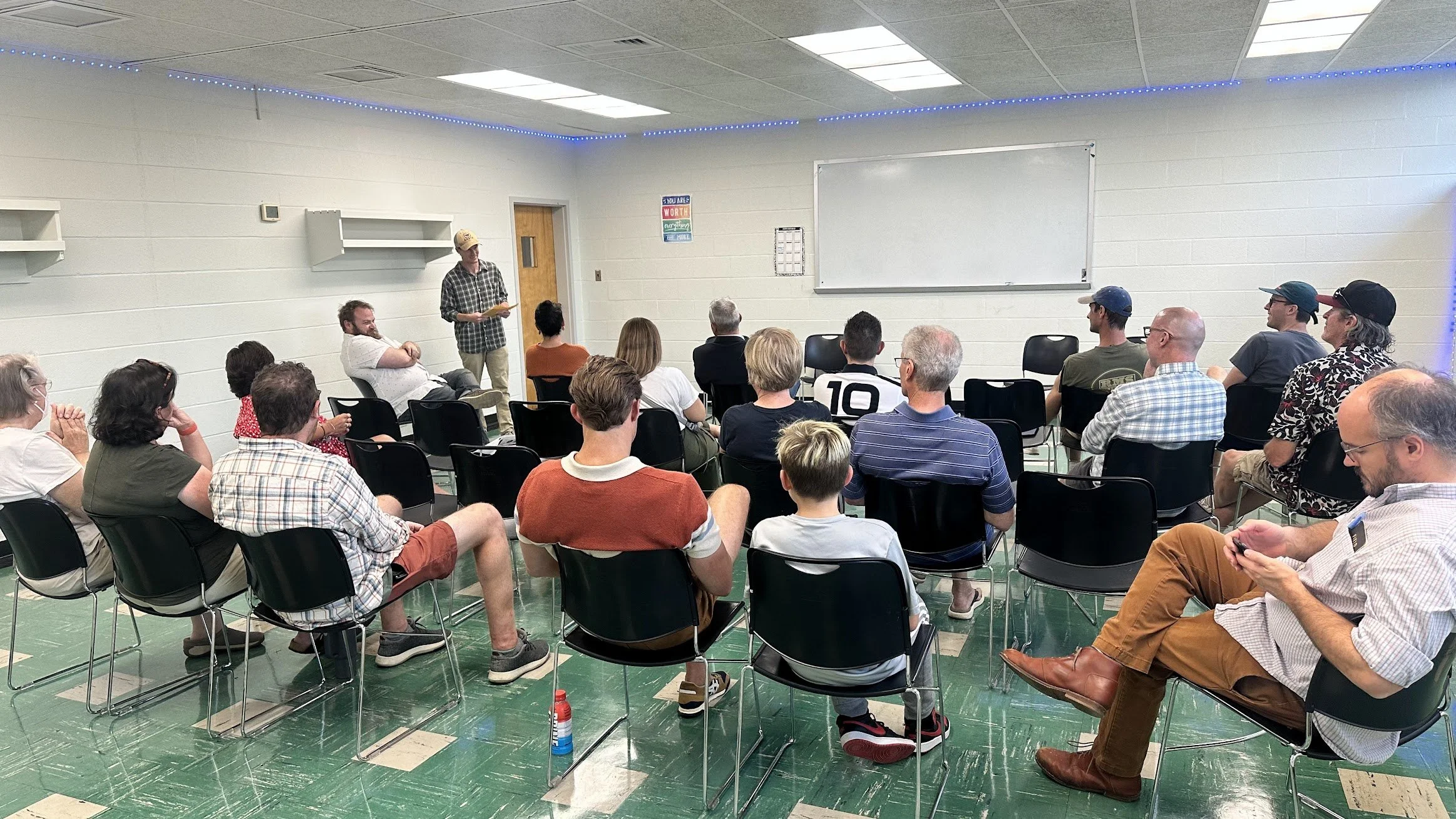 A diverse group of people attending a meeting or presentation in a small room with white walls and a tiled green floor. One person is standing at the front, speaking or presenting, while others are seated and listening. The room has ceiling lights, wall shelves, and warnings or motivational signs on the wall.