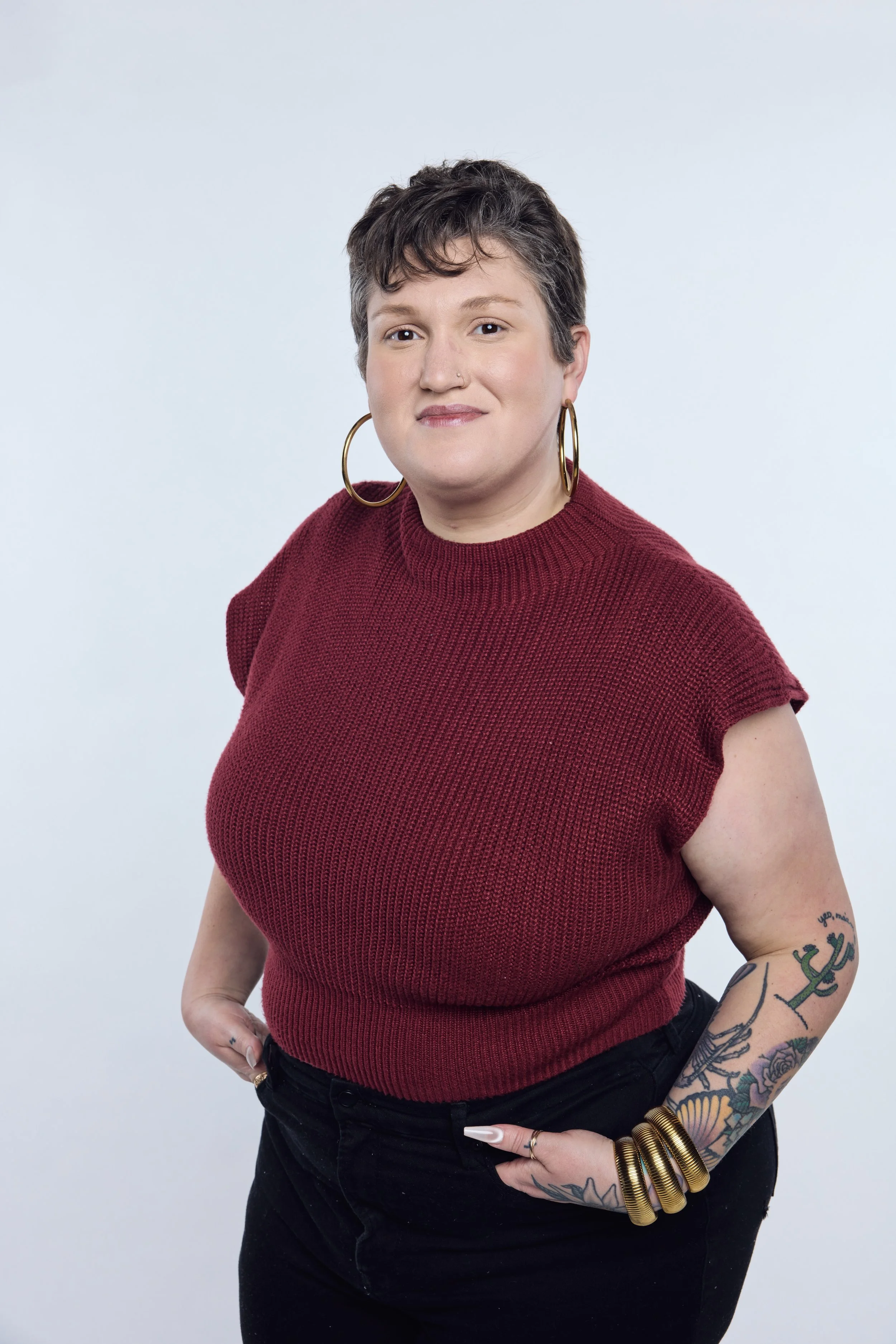 A woman with short dark hair and large hoop earrings, wearing a red knitted top and black pants, standing against a plain light background.