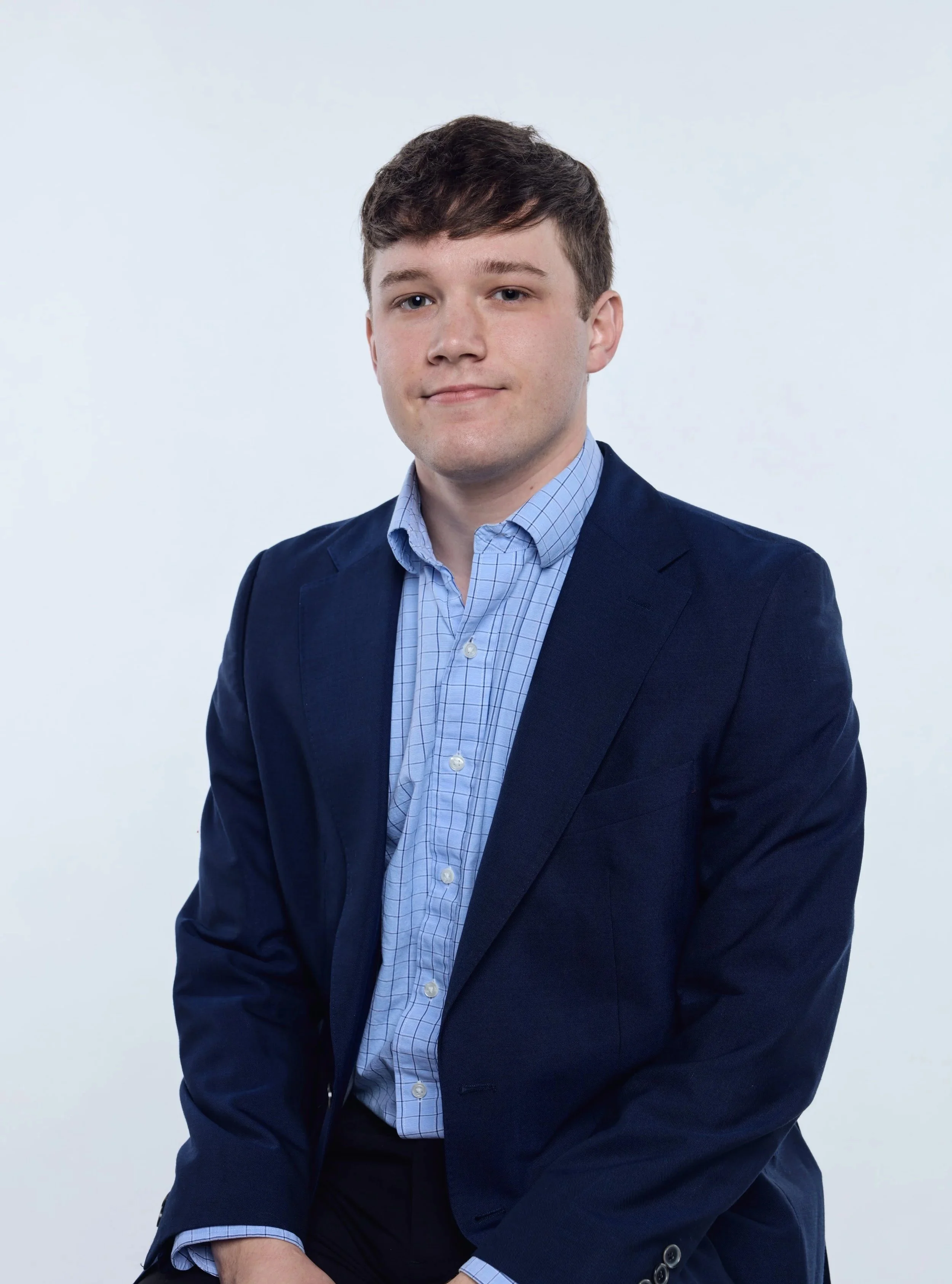 Young man in navy blazer and light blue checked shirt, posing against a plain white background, looking directly at the camera with a slight smile.