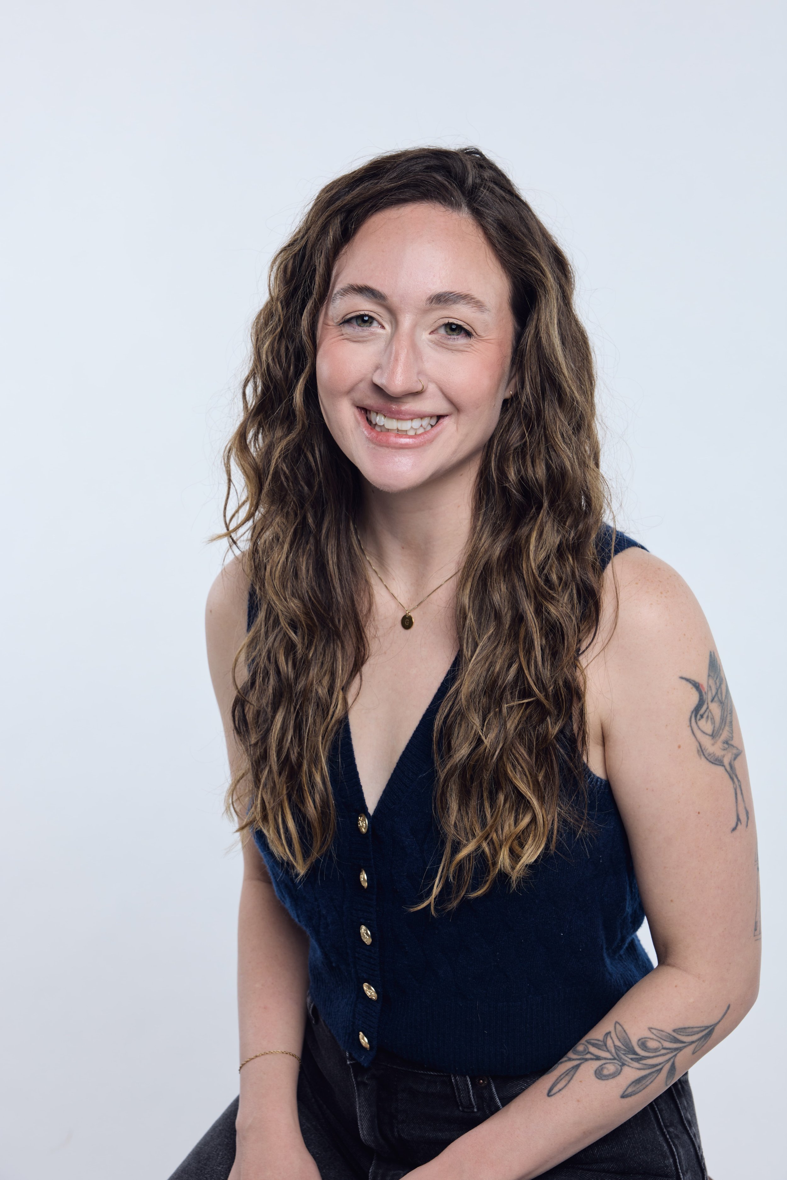 A woman with long wavy brown hair smiling, wearing a dark sleeveless top with gold buttons, a gold necklace, and tattoos on her left arm.