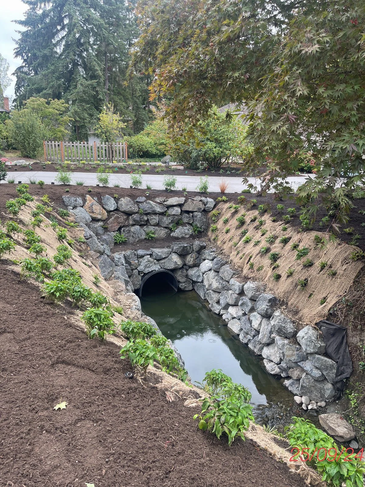 Photograph of a newly constructed drainage culvert with stone walls and a water channel flowing through it, surrounded by landscaped soil and young plants.