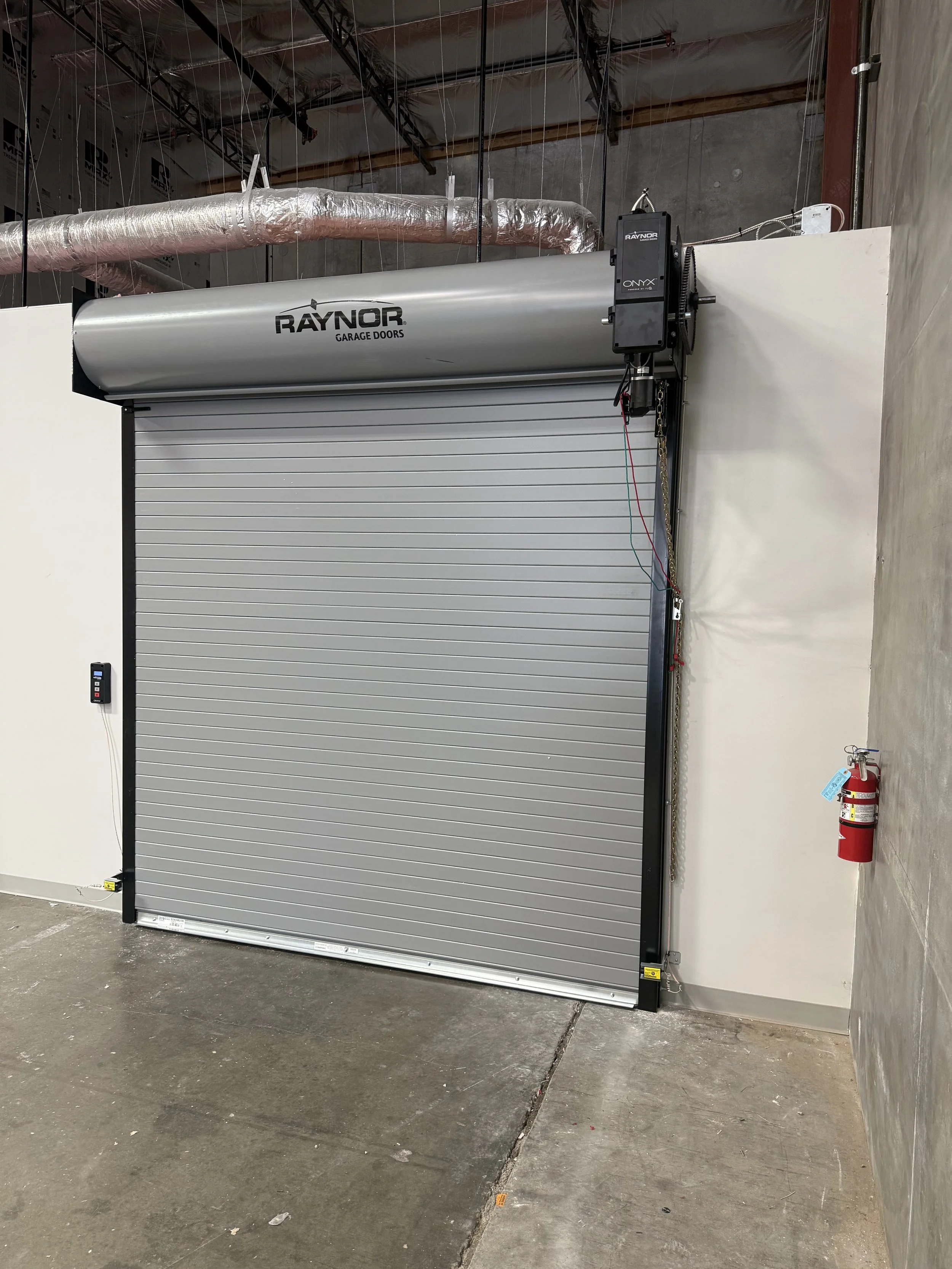 A partially open garage door with the RAYNOR brand logo, installed in a warehouse or workshop setting, with a fire extinguisher and a remote control unit nearby.