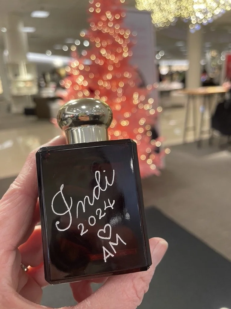 A hand holding a perfume bottle with a silver cap, engraved with "Jade 2024 ❤️ AM". In the background, blurred Christmas tree with red decorations and colorful lights.