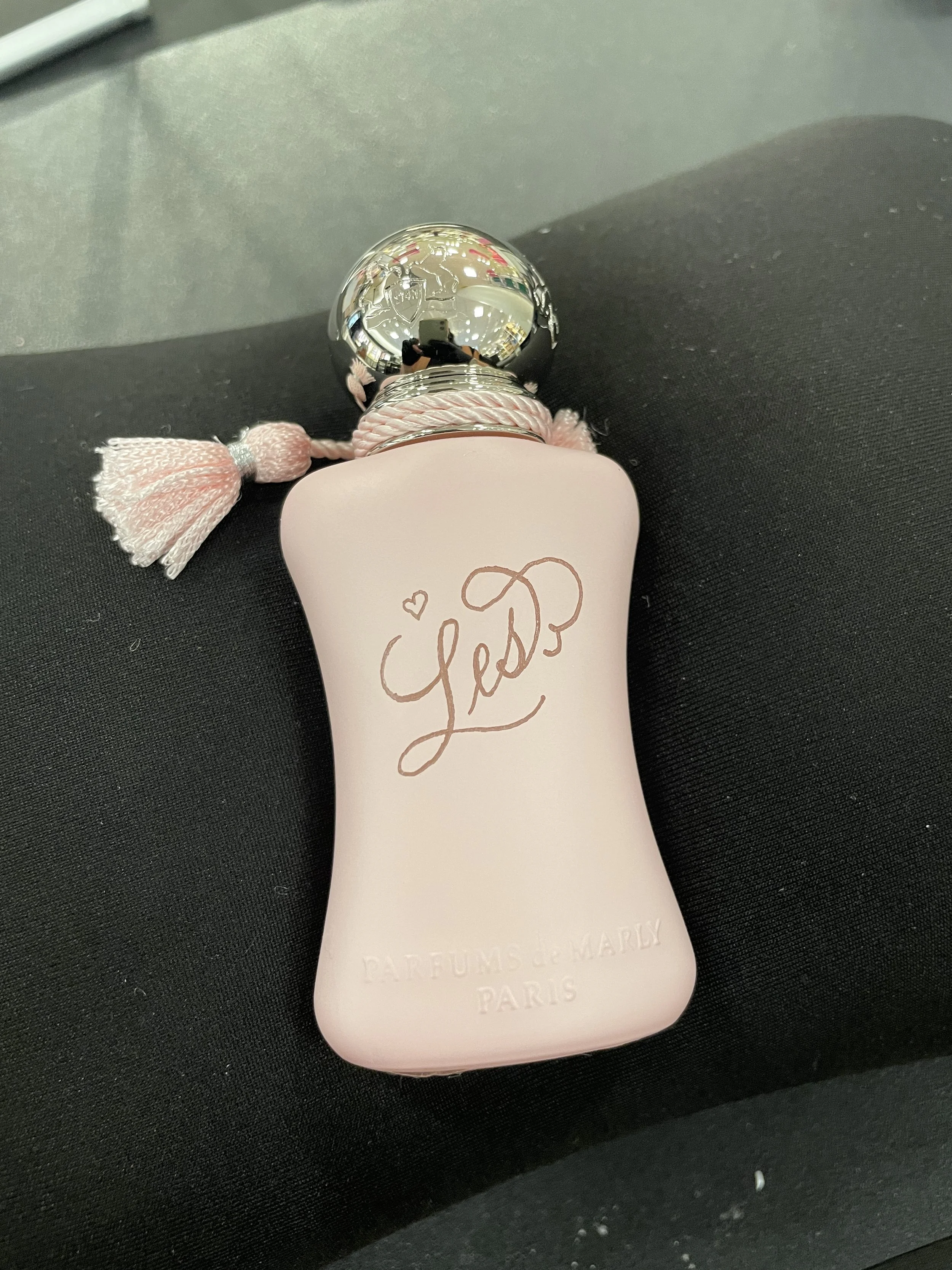 A bottle of Love perfume from Parfums de Marly, Paris, has a pastel pink body with a shiny silver spherical cap and a tassel attached to the neck, resting on a black fabric surface.
