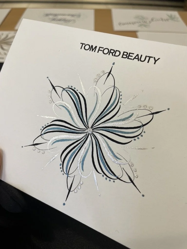 Front of a white card with 'TOM FORD BEAUTY' printed at the top and an intricate floral design in black, white, silver, and blue. The card is being held by a person in a dimly lit room.