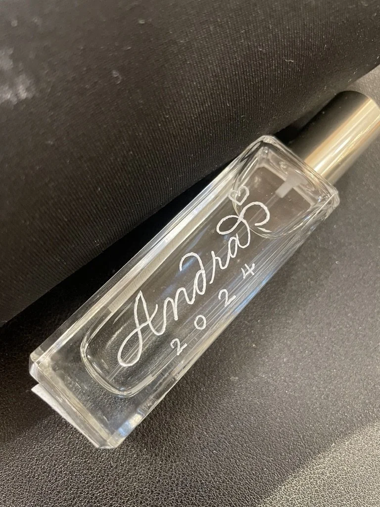 A clear glass perfume bottle with silver cap, engraved with the name 'Amanda' and the date '2024.2.14', placed on a black textured surface.