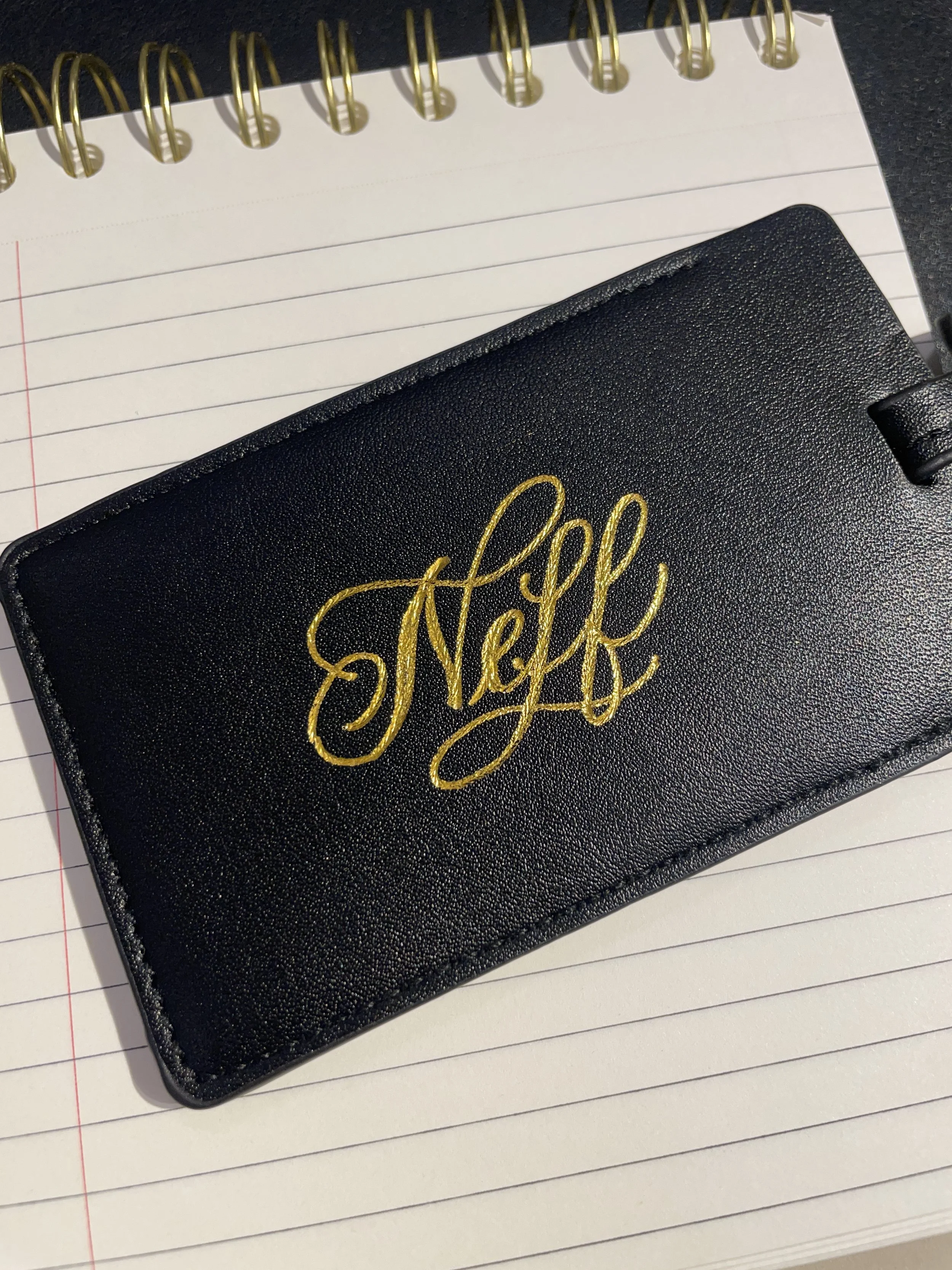 A black wallet with gold embroidered text that reads 'Nek' resting on an open, spiral-bound notebook with lined pages.