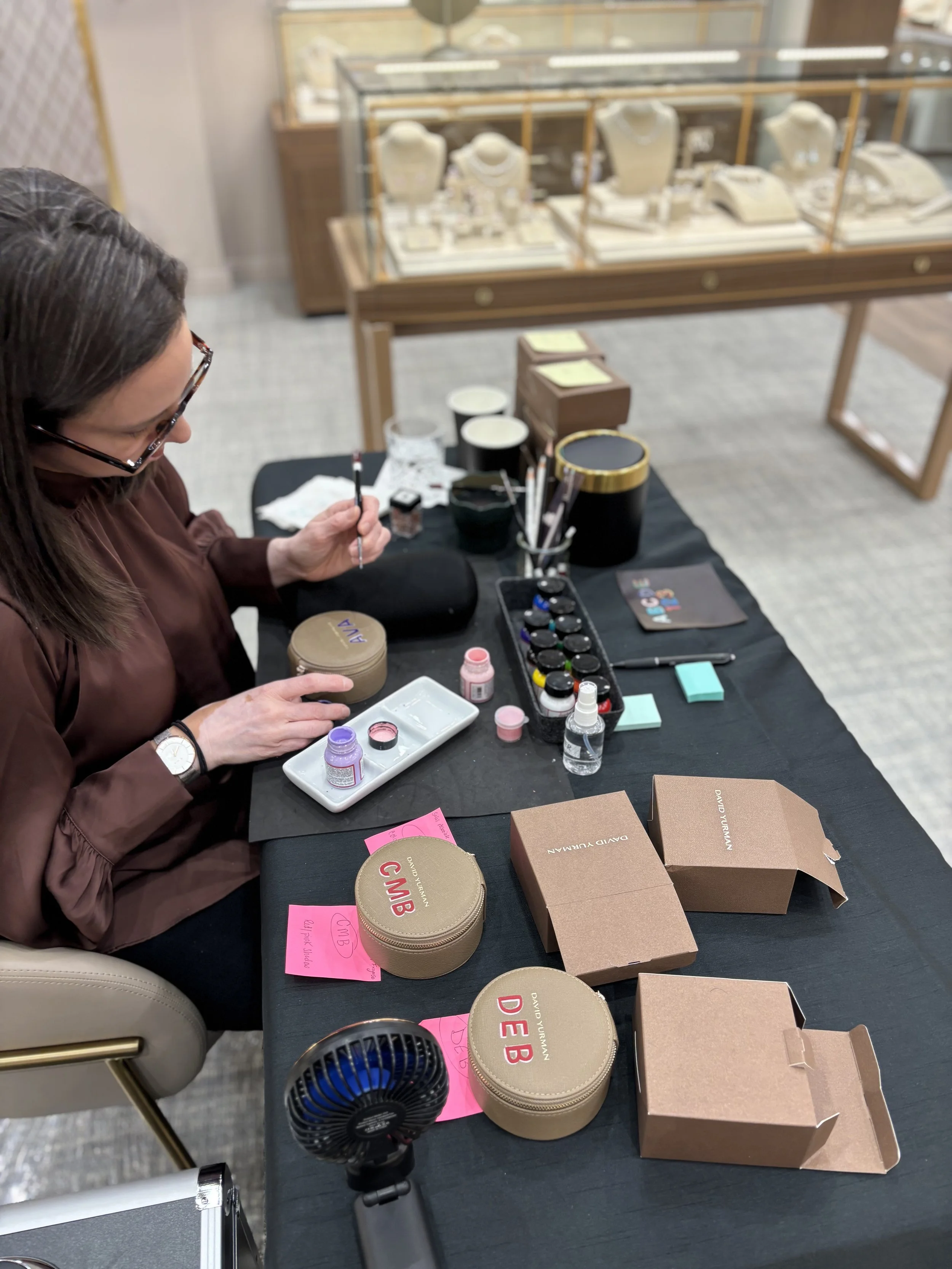 A woman sitting at a table, working with leather paints, with various containers and boxes on the table. In the background, there are jewelry display cases with necklaces and other jewelry pieces. James Free Jewelers