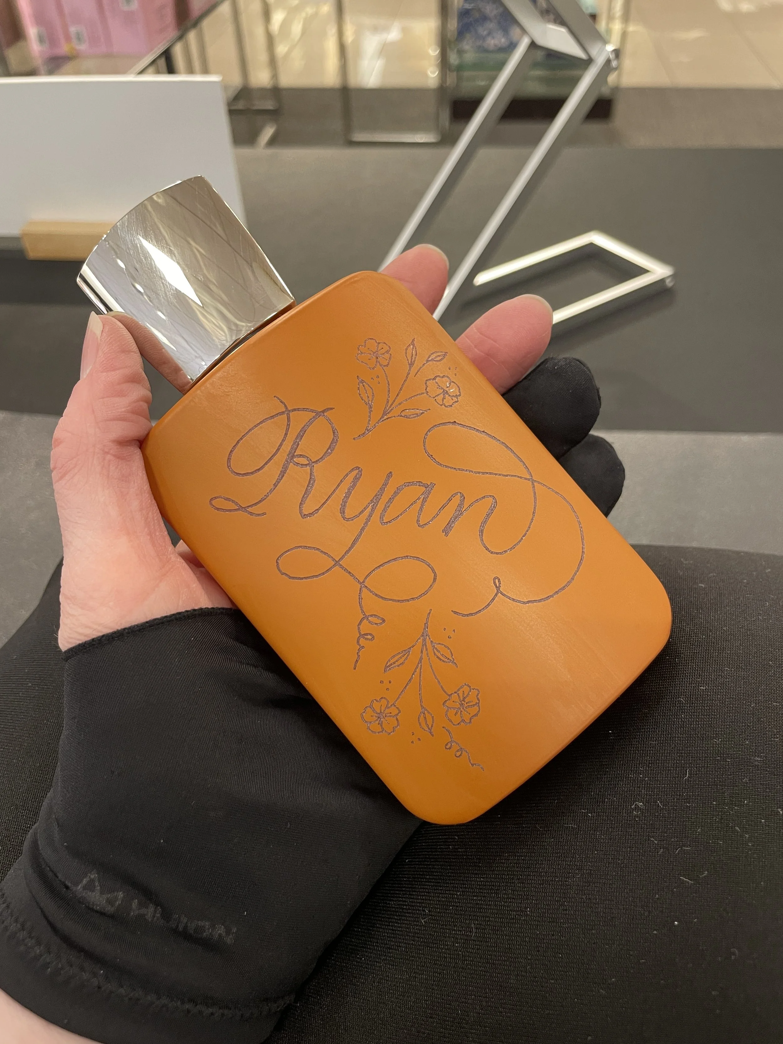Orange bottle with a silver cap, decorated with handwritten-style text that says 'Ryan' and floral designs, held in a person's hand wearing a black glove.
