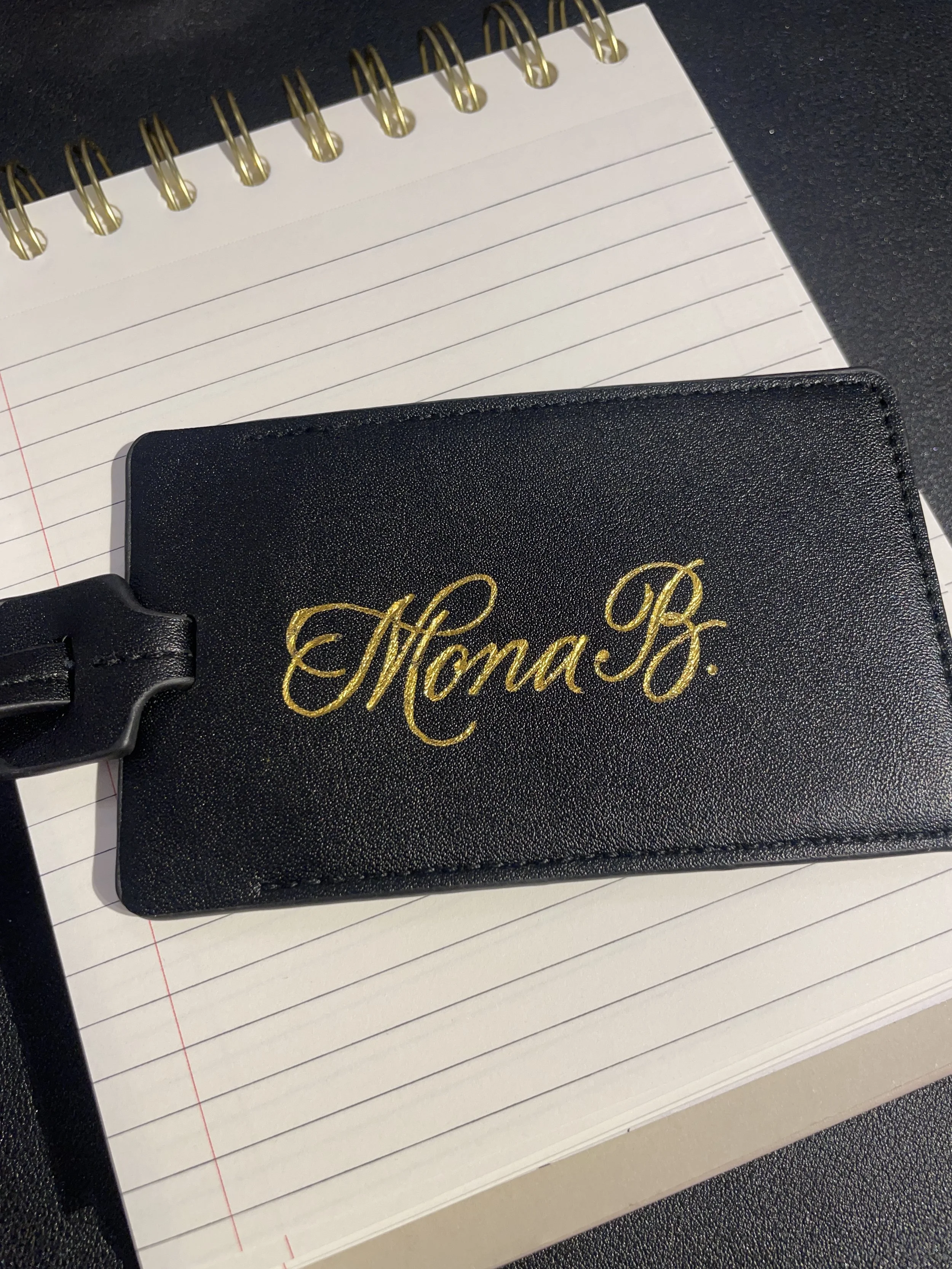 A black leather luggage tag with gold embossed cursive initials resting on a lined notebook with a spiral binding, on a dark surface.