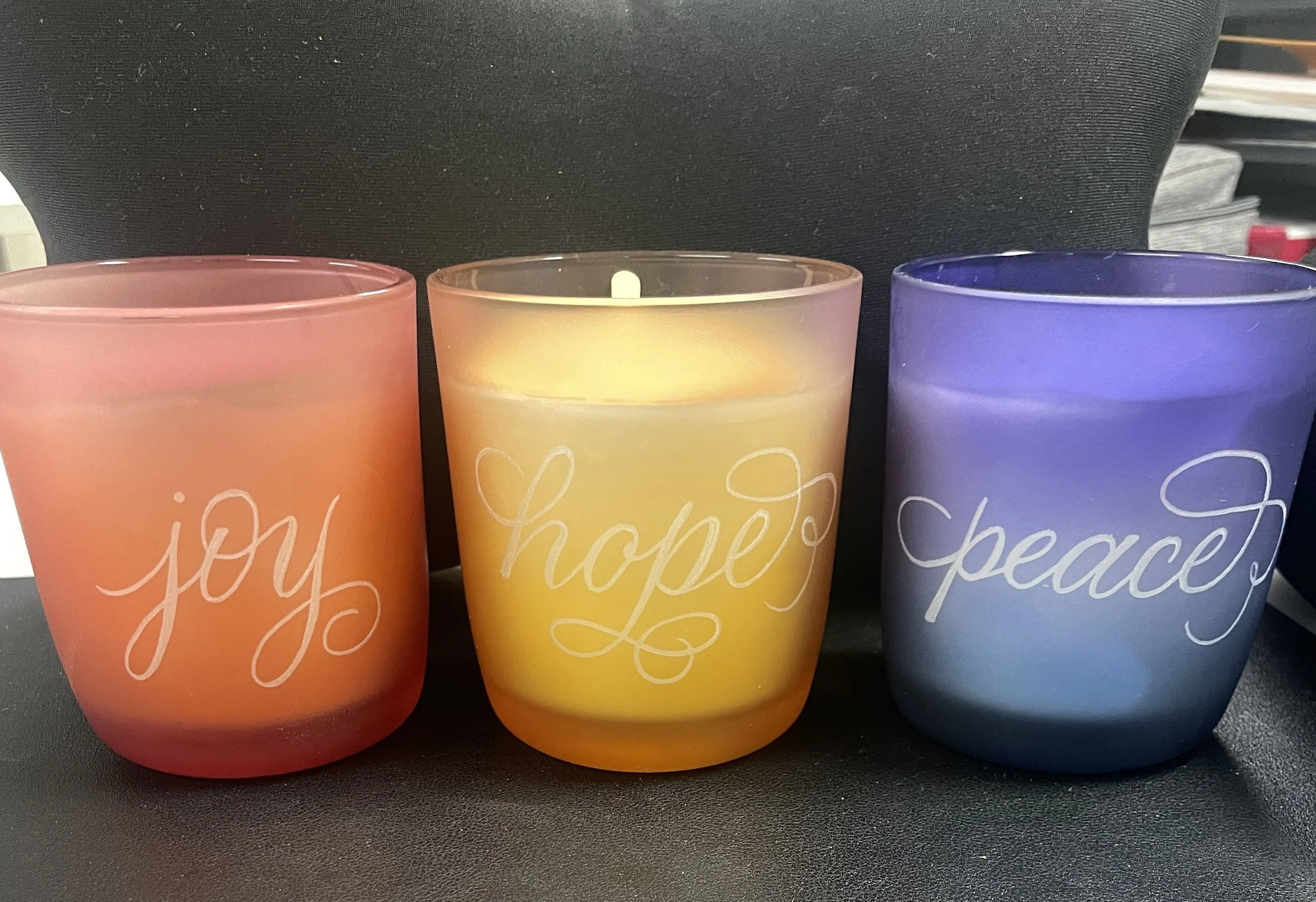 Three frosted glass candles in pink, yellow, and purple, each with words 'joy', 'hope', and 'peace' engraved on them, placed on a black surface.