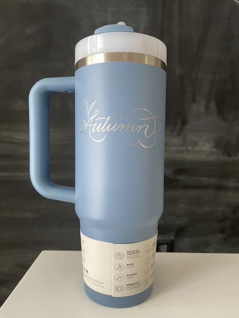 A light blue travel mug with the word 'Autumn' engraved in cursive on it, placed on a white surface against a dark background.
