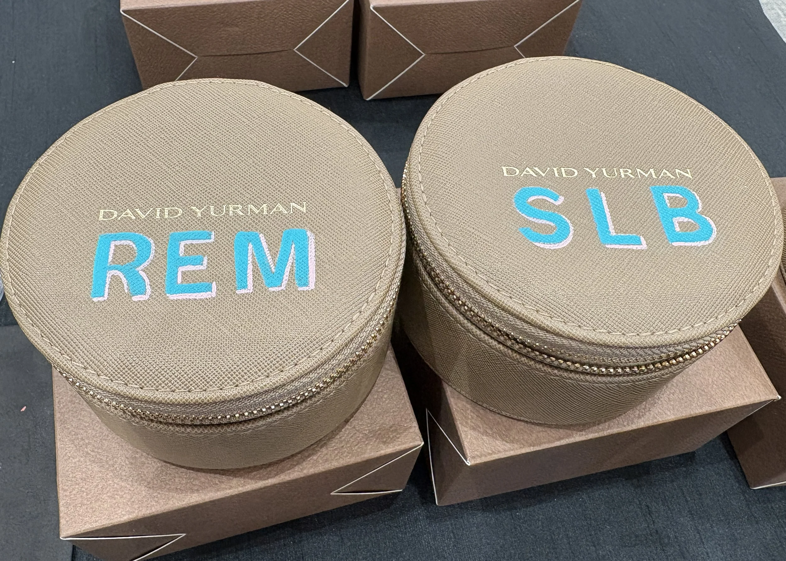 Round beige cases with embroidered text 'DAVID YURMAN' and 'REM' on one, and 'SLB' on the other, placed on top of brown boxes. Leather Painting