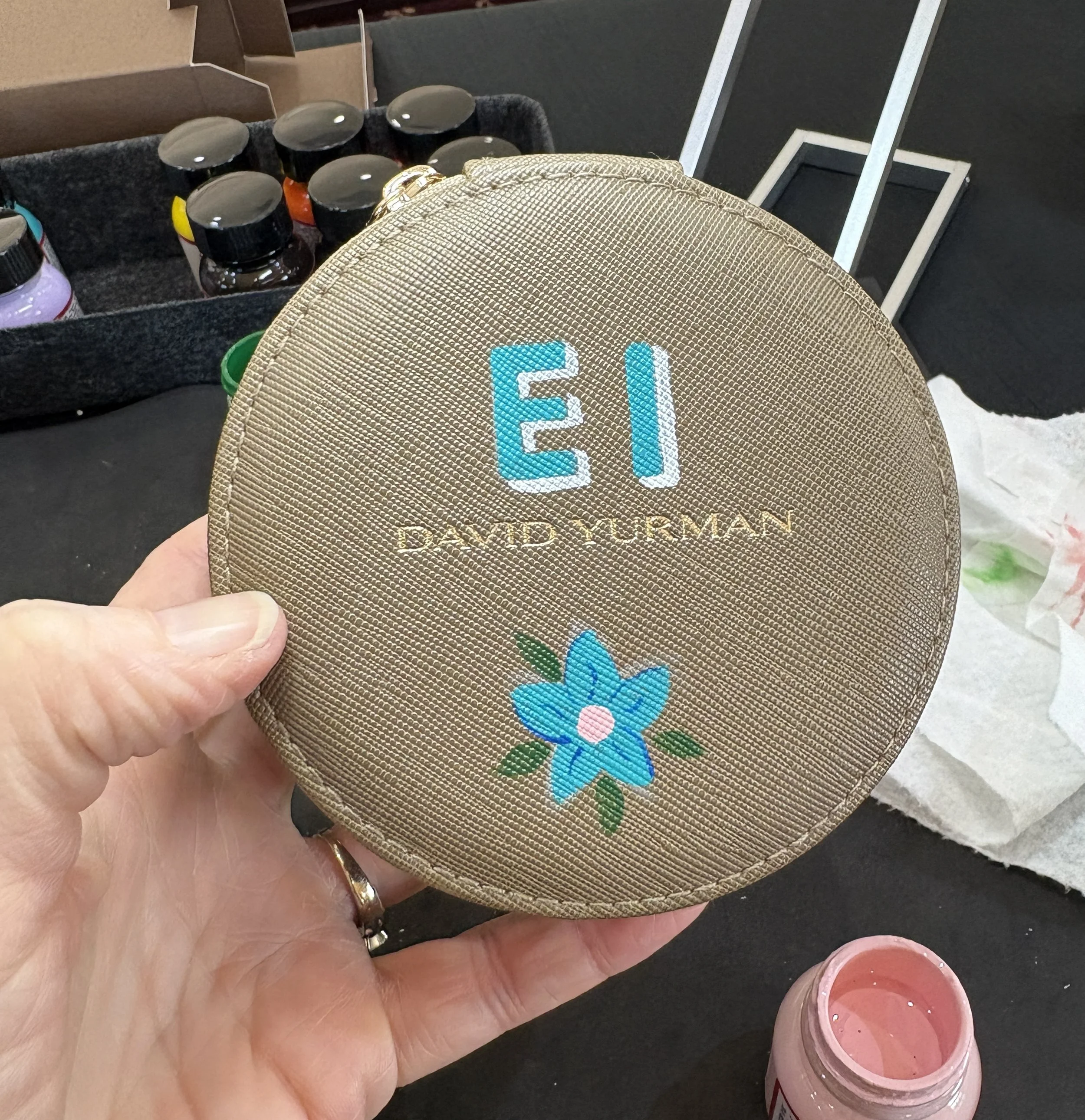 A round tan-colored canvas pouch with the name 'Eli' embroidered in blue letters, 'David Yurman' written in gold, and a blue flower with green leaves on it, held by a person's left hand.