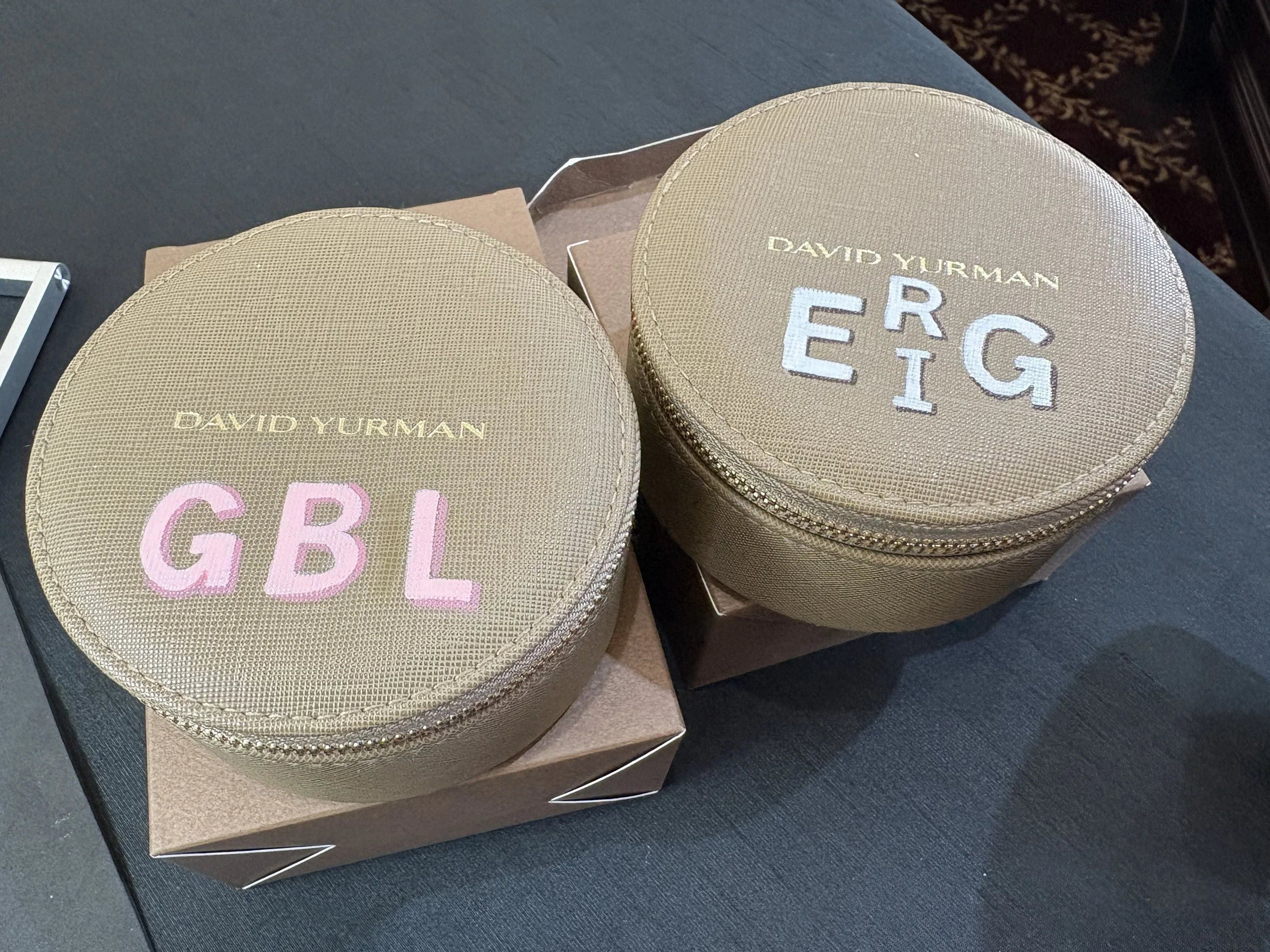 Two round beige leather jewelry boxes painted 'G.B.L' in pink and 'E.G' in blue, with the brand name 'David Yurman' on top, placed on top of two brown boxes on a dark surface.