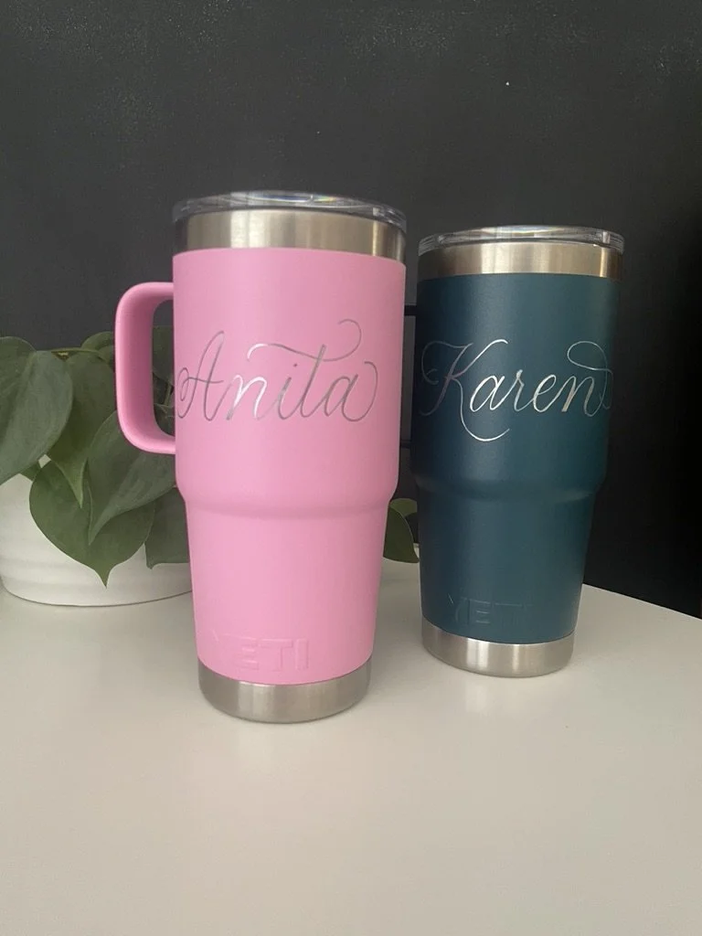 Two stainless steel travel mugs, one pink with the name 'Anita' and one teal with the name 'Karen,' placed on a white surface next to a green leafy plant.
