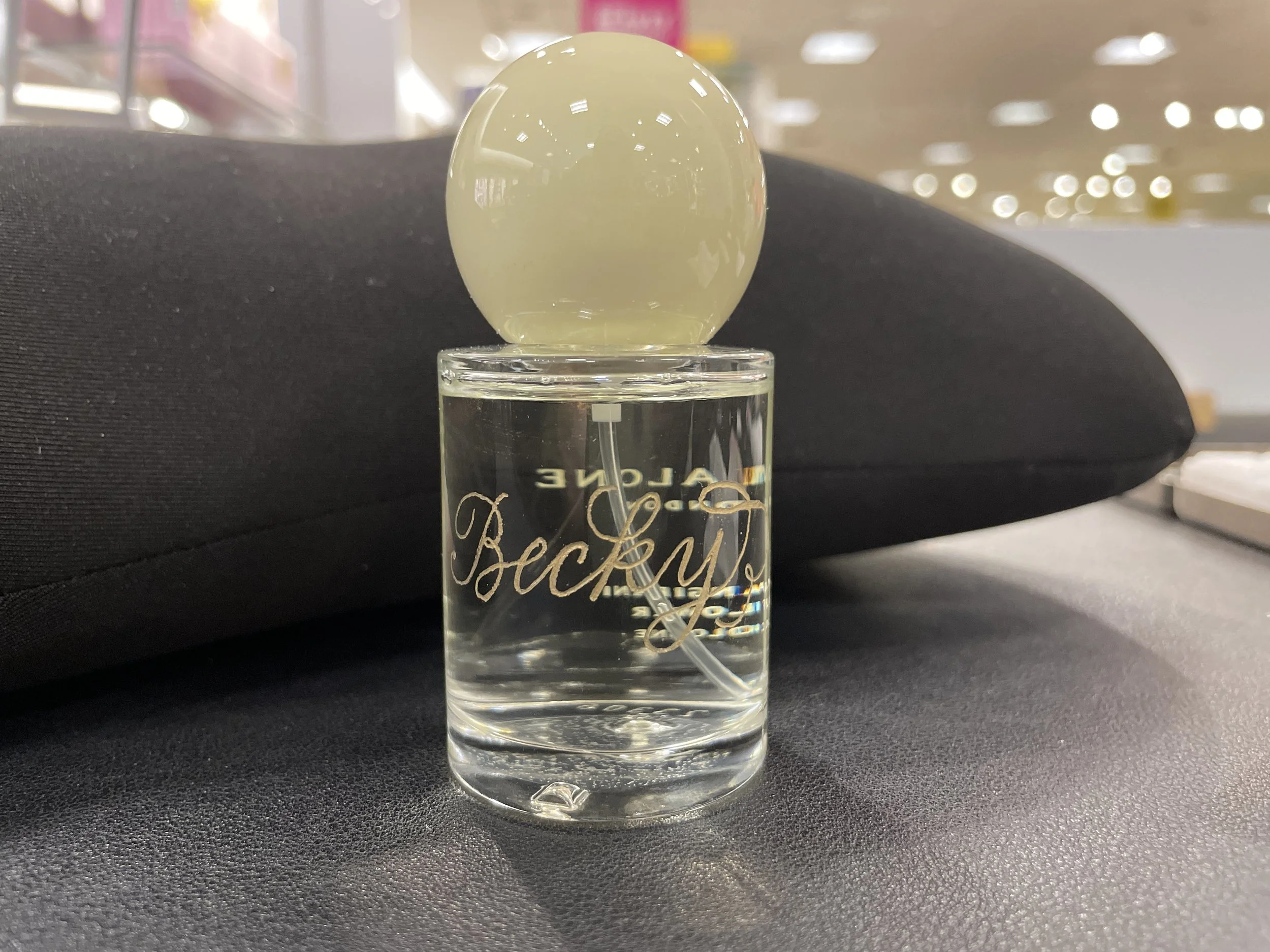 A perfume bottle with a rounded, pale yellow-green cap is placed on a dark surface. The bottle has the brand name 'Baccarat' engraved on it and is filled with a clear liquid. The background shows a blurred retail store interior with bright lighting.