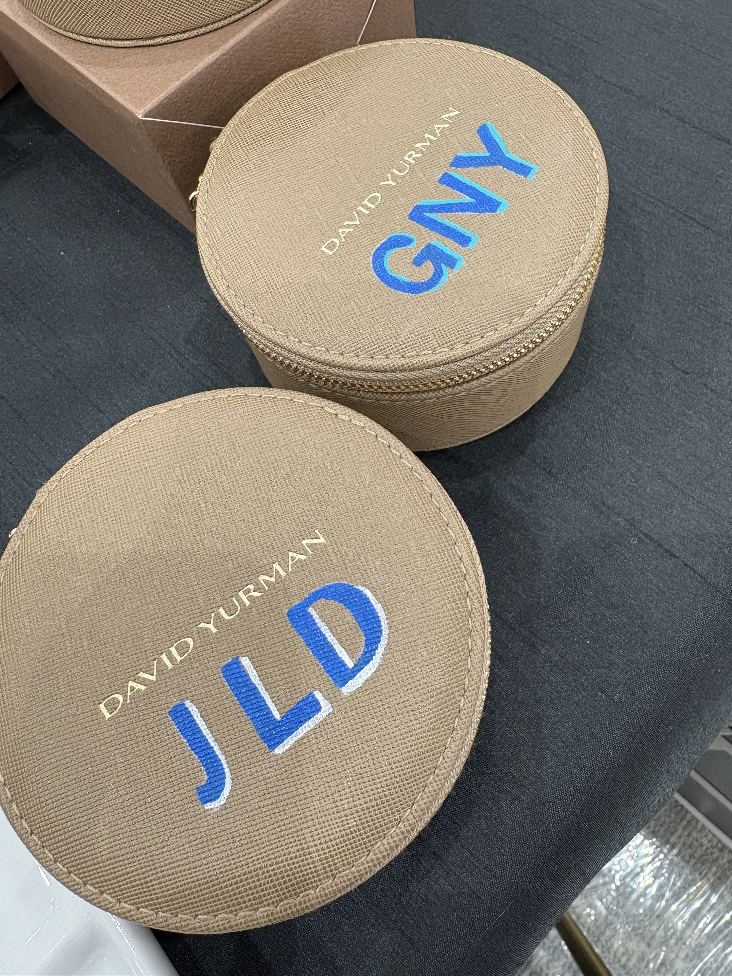 Two beige round cases with blue embroidered initials 'JLD' and the brand name 'David Yurman' on the top case, placed on a black surface.