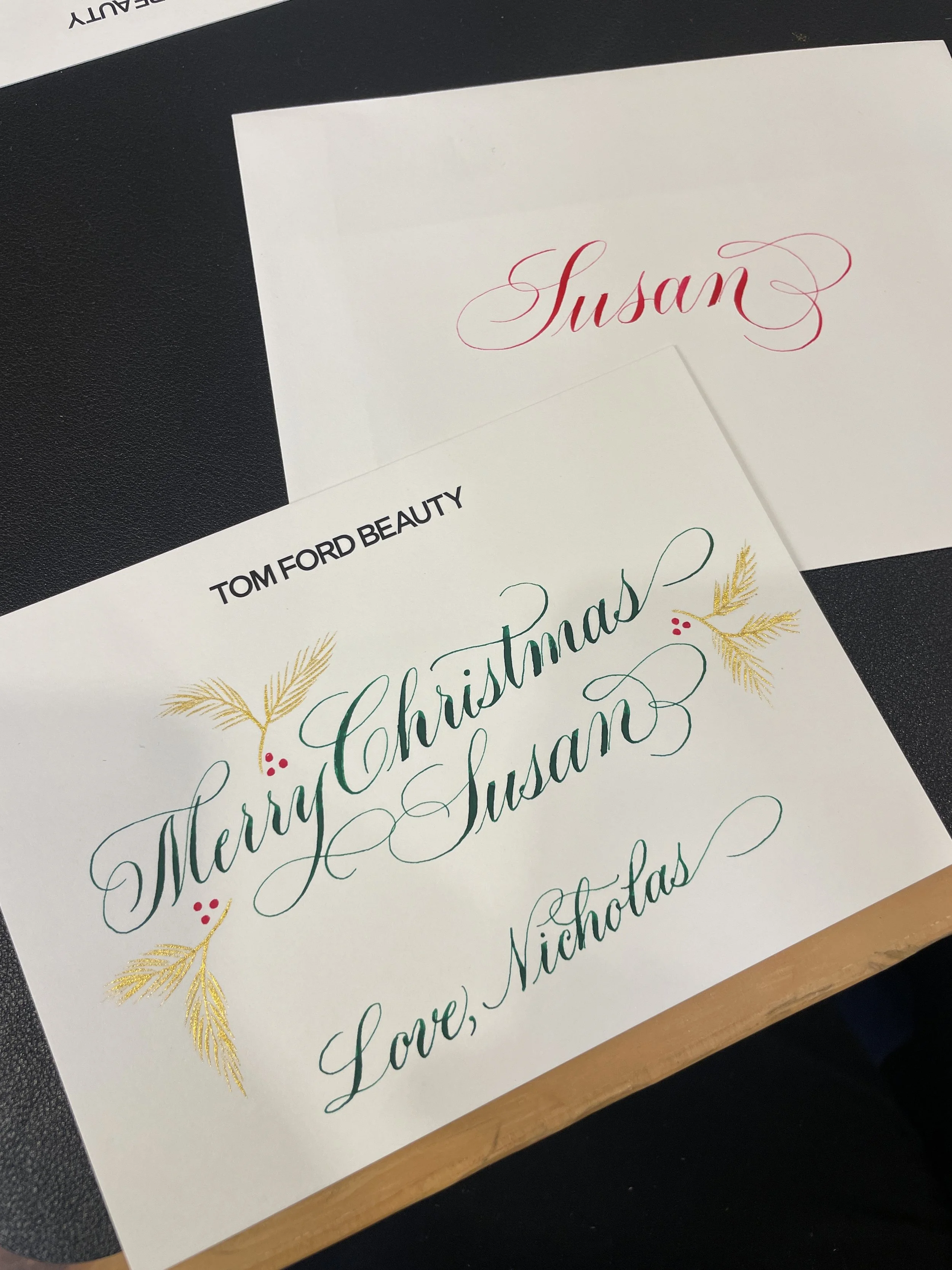 A Christmas greeting card with green and gold lettering, decorated with gold pine branches and red berries, reading "Merry Christmas Susan Love, Nicholas." Another card in the background has the name "Susan" written in red cursive.